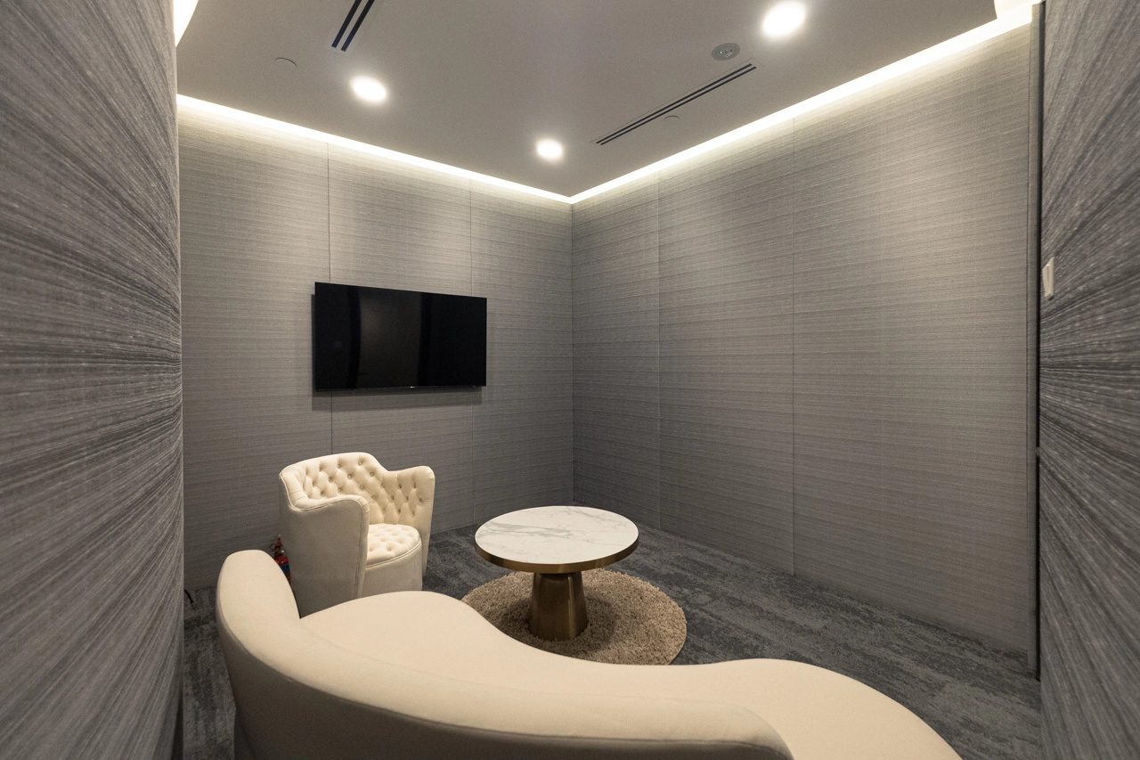 A small, modern lounge room with gray walls, a wall-mounted flat screen TV, a round marble coffee table on a circular rug, a white tufted chair, and a curved white sofa under recessed ceiling lights.