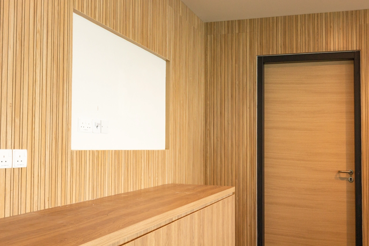 A modern interior with light wood panel walls, a wooden door with a dark frame, a wooden countertop, power outlets, and a large white rectangular cutout in the wall above the counter.