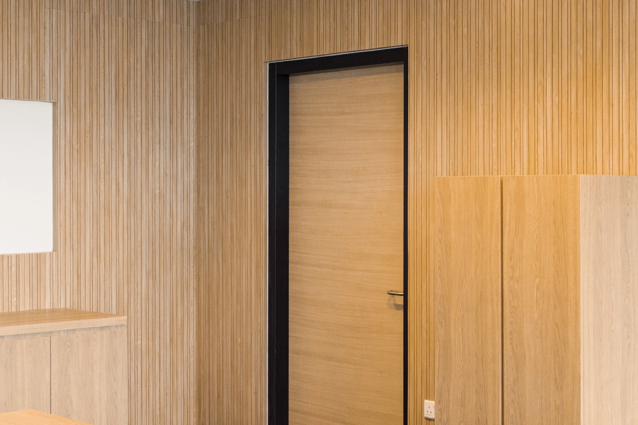 A modern room with light wood panel walls, a closed wooden door with a black frame, a white rectangular wall panel, and a wooden cabinet.