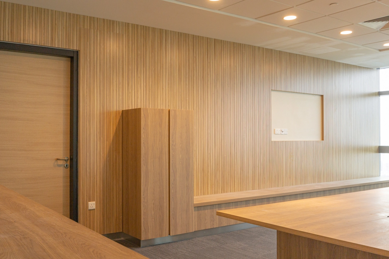 A modern office with light wood panel walls, wooden furniture, a closed wooden door, a tall cabinet, a long bench, carpeted floor, and a window letting in natural light.