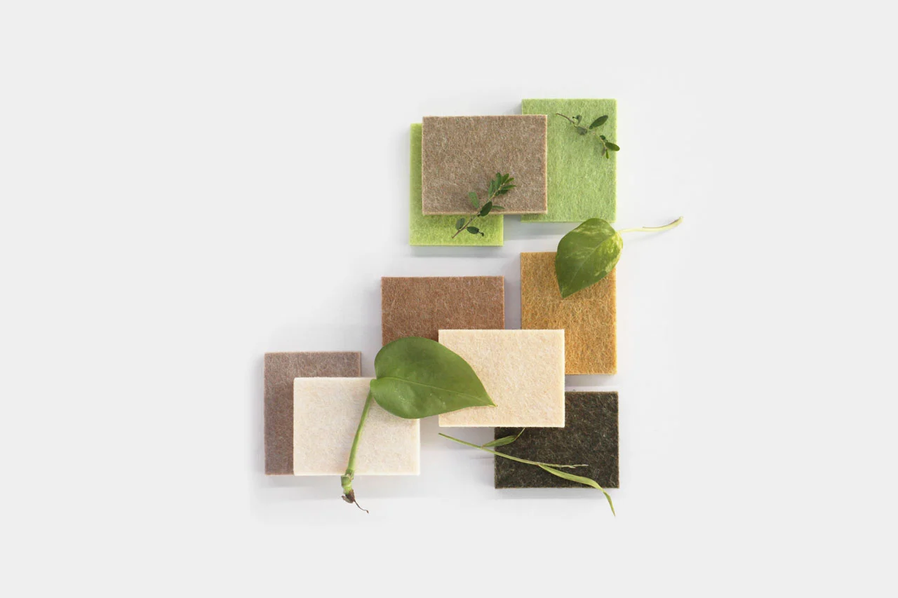 A collection of rectangular fabric samples in earth tones such as brown, beige, green, and cream, with green plant leaves and tiny vines placed artfully on top. The samples are stacked on a light grey background.