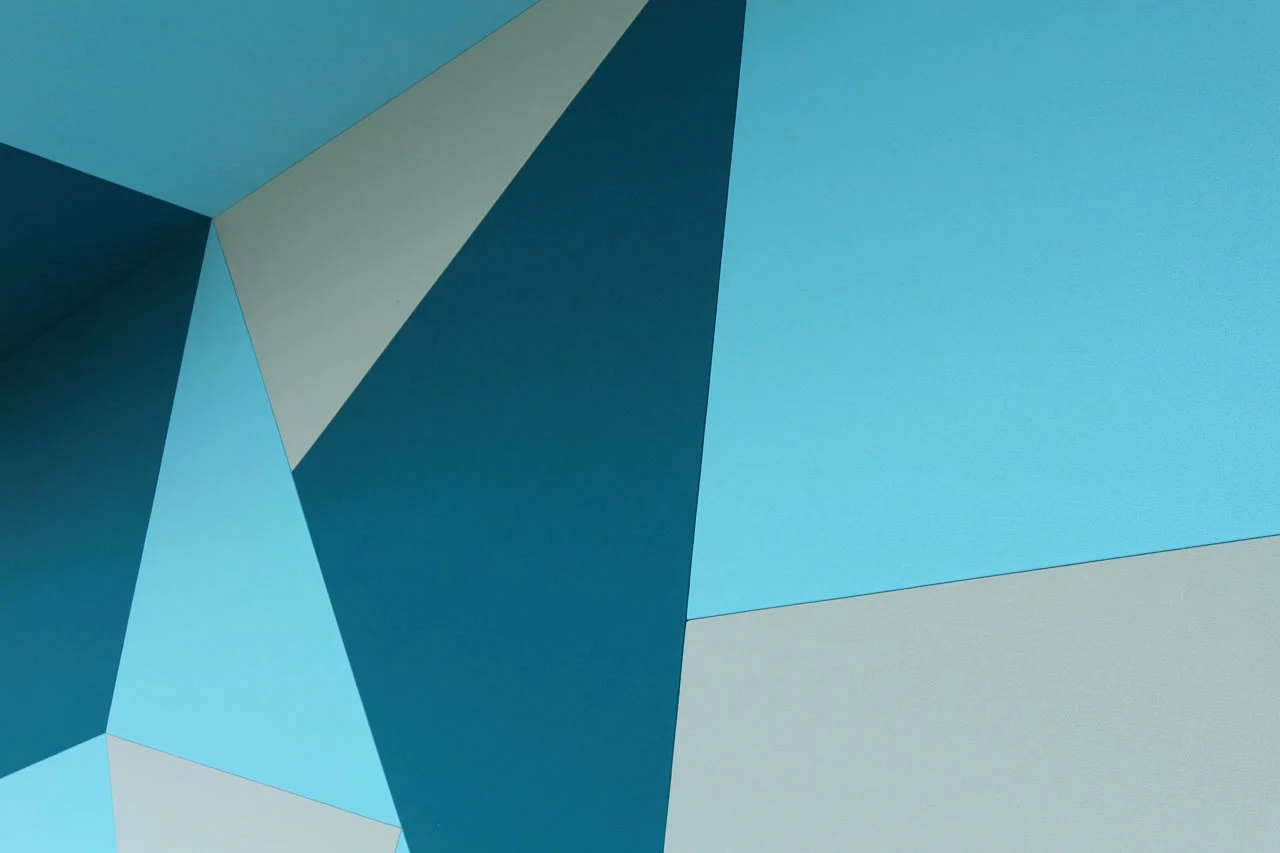 Abstract geometric pattern featuring overlapping shapes in various shades of blue and light gray. The design creates a dynamic, modern look with sharp angles and contrasting colors.