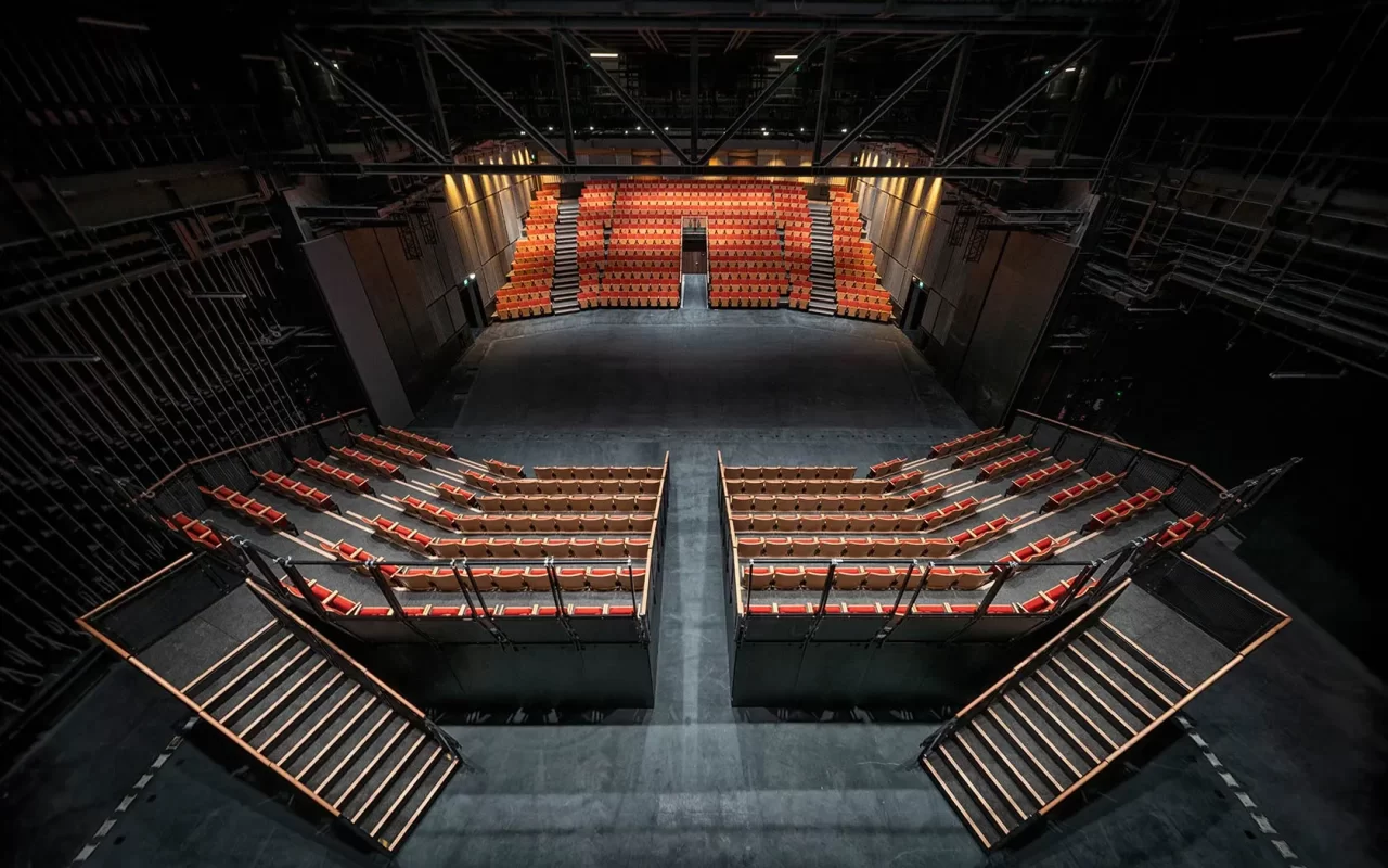 A view from the stage of an empty, modern theater with orange seats arranged in sections, staircases on both sides, and a large open space between the stage and the audience.