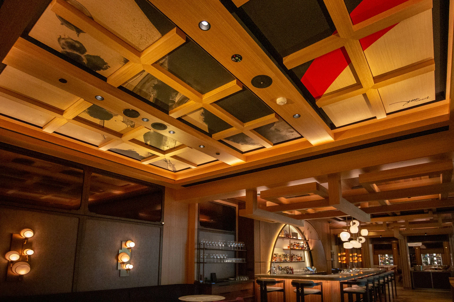 A modern bar with wooden lattice ceiling, warm lighting, a row of bar stools, shelves with glasses and bottles, and unique wall light fixtures creating a cozy atmosphere.