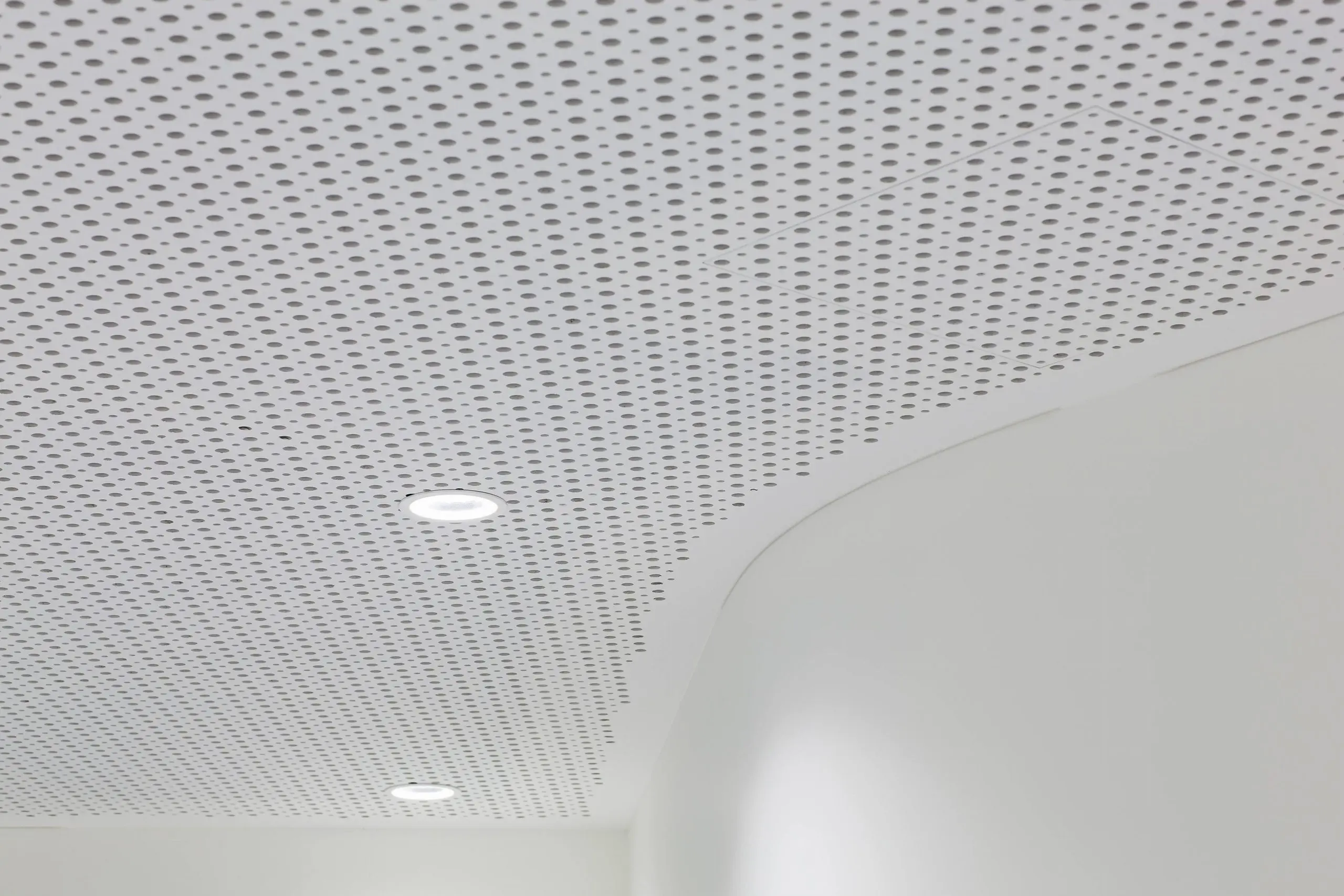 A white perforated ceiling with two recessed round lights and a curved wall below. The ceiling features a pattern of small, evenly spaced holes. The space appears modern and minimalistic.