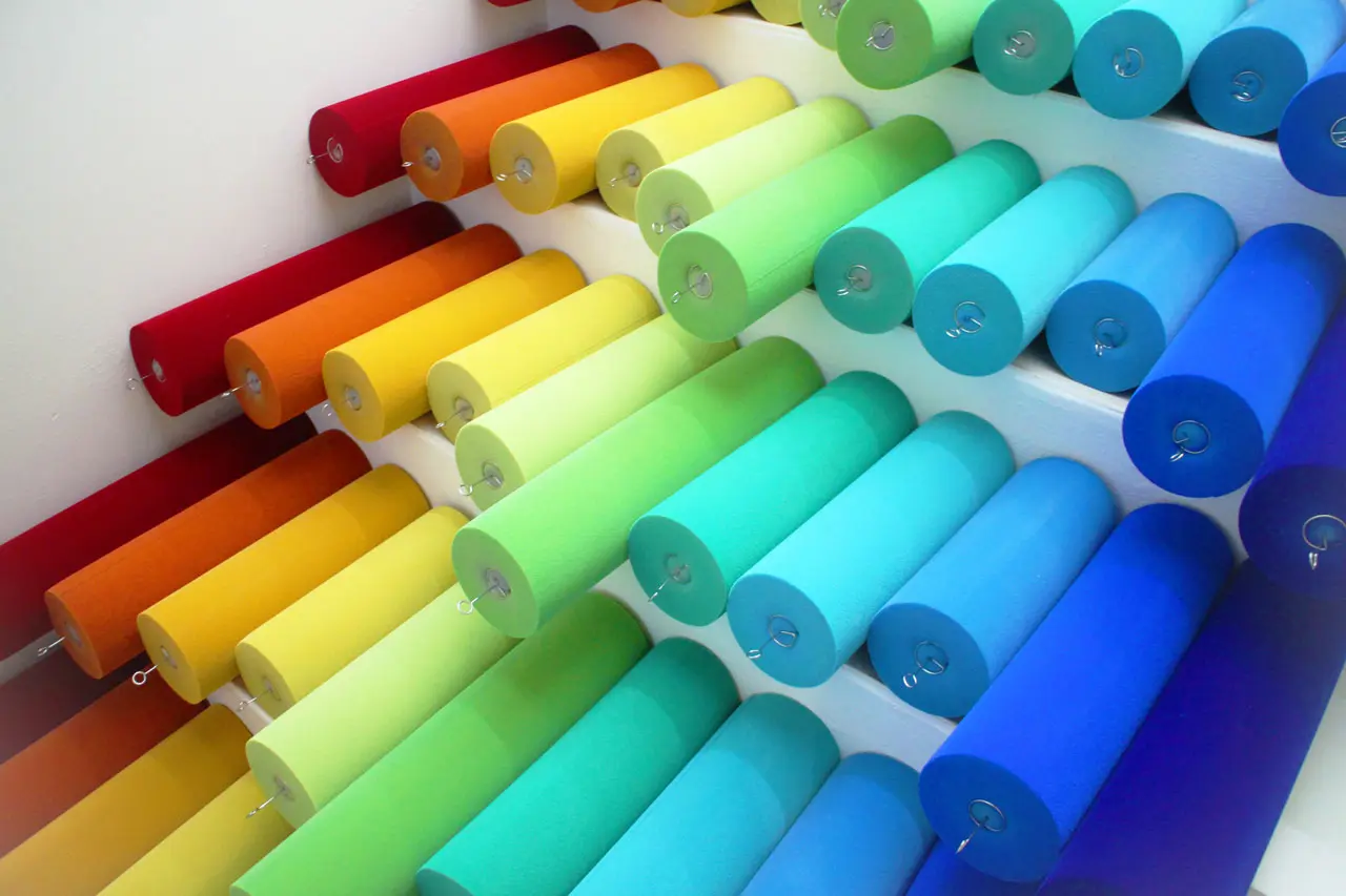 Rows of large fabric rolls are neatly arranged on slanted shelves, showcasing a gradient of colors from red, orange, yellow, and green to blue and turquoise. The display creates a vibrant and orderly color spectrum.