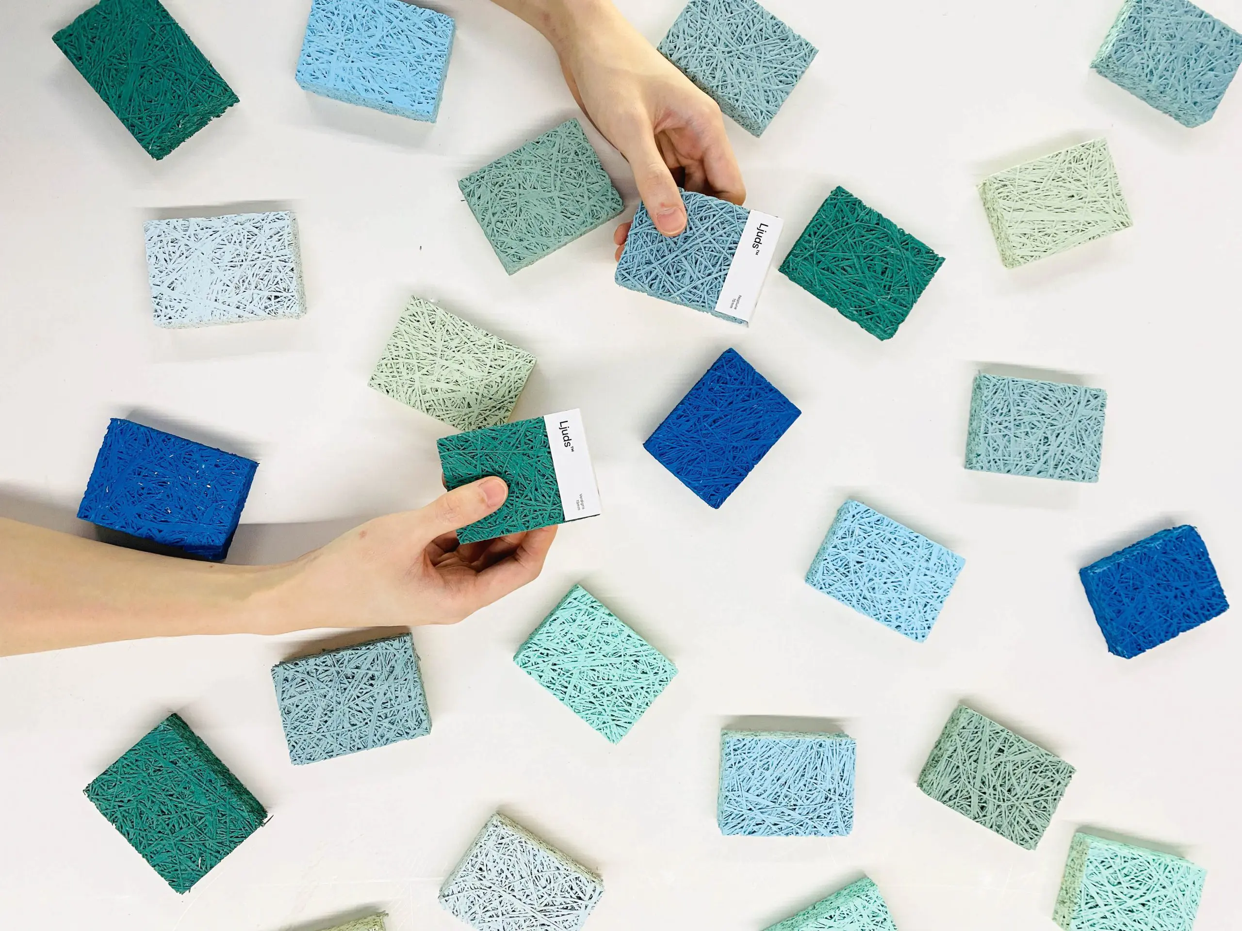 A close-up view of two hands selecting from multiple textured sample blocks in shades of blue and green. The blocks are placed randomly on a white surface.