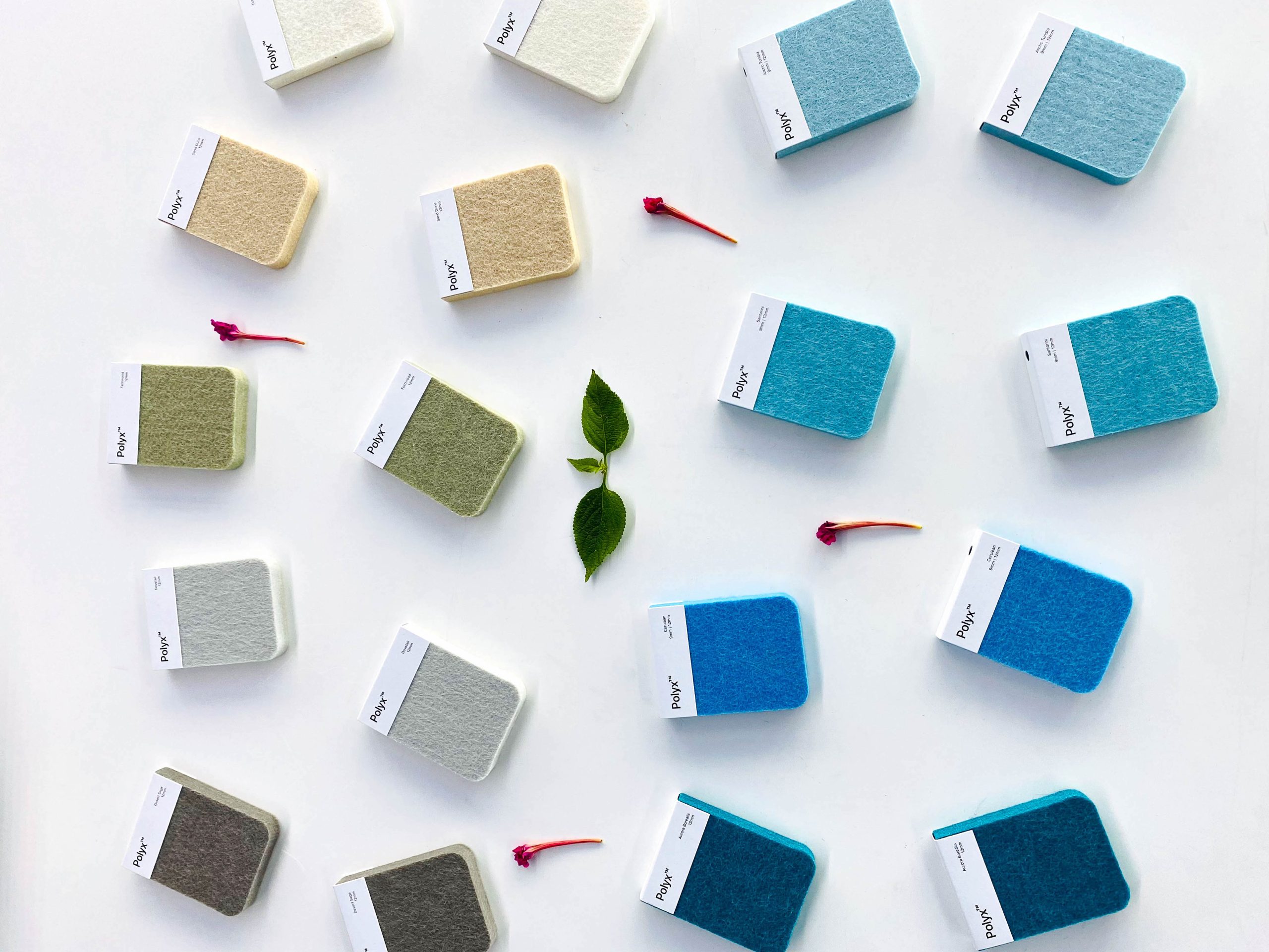 A neat arrangement of fabric sample squares in light colors such as white, beige, and shades of blue and green, laid out on a white background. Small leaves and pink flower petals are scattered between the samples.