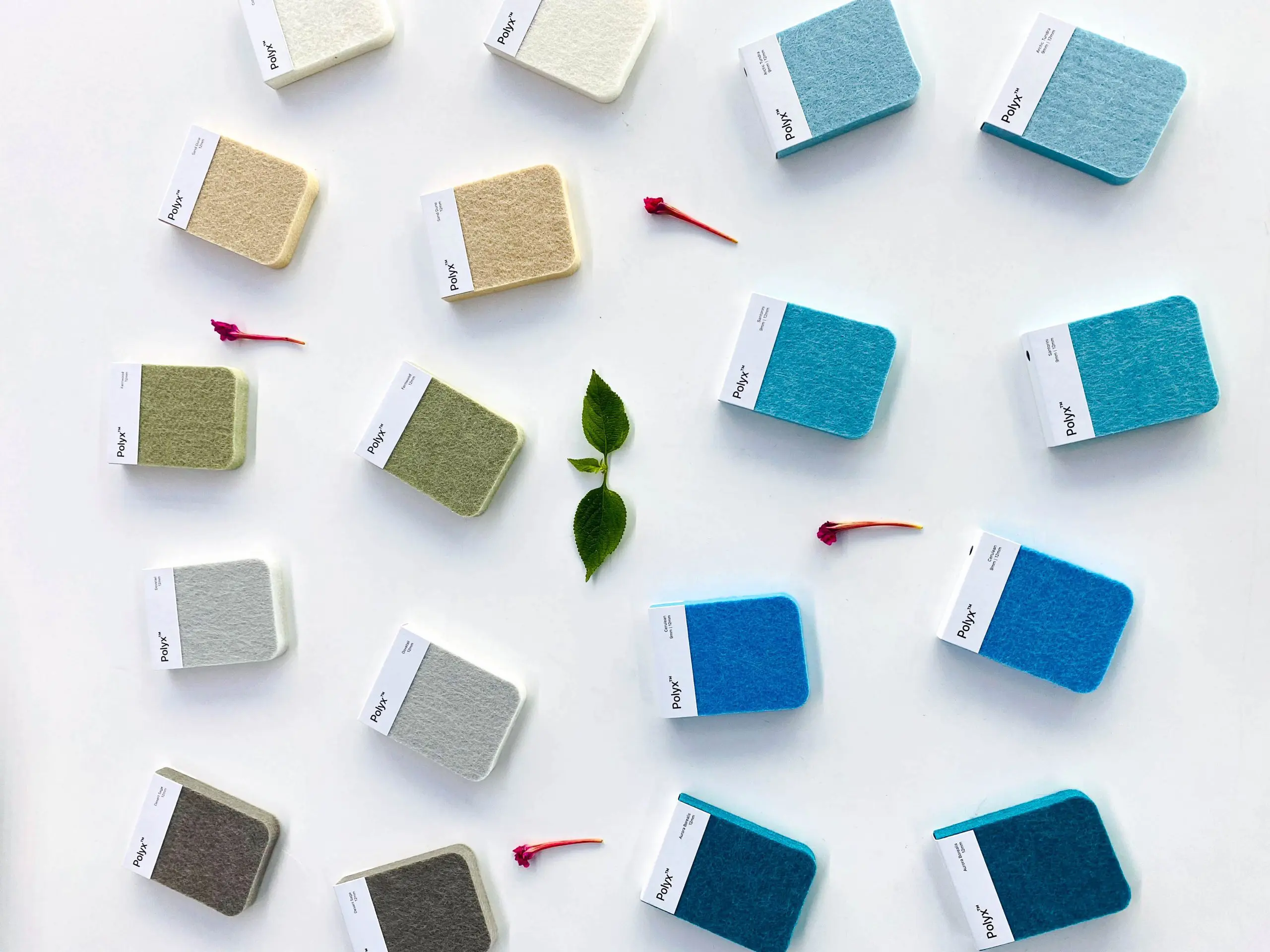 A neat arrangement of fabric sample squares in light colors such as white, beige, and shades of blue and green, laid out on a white background. Small leaves and pink flower petals are scattered between the samples.