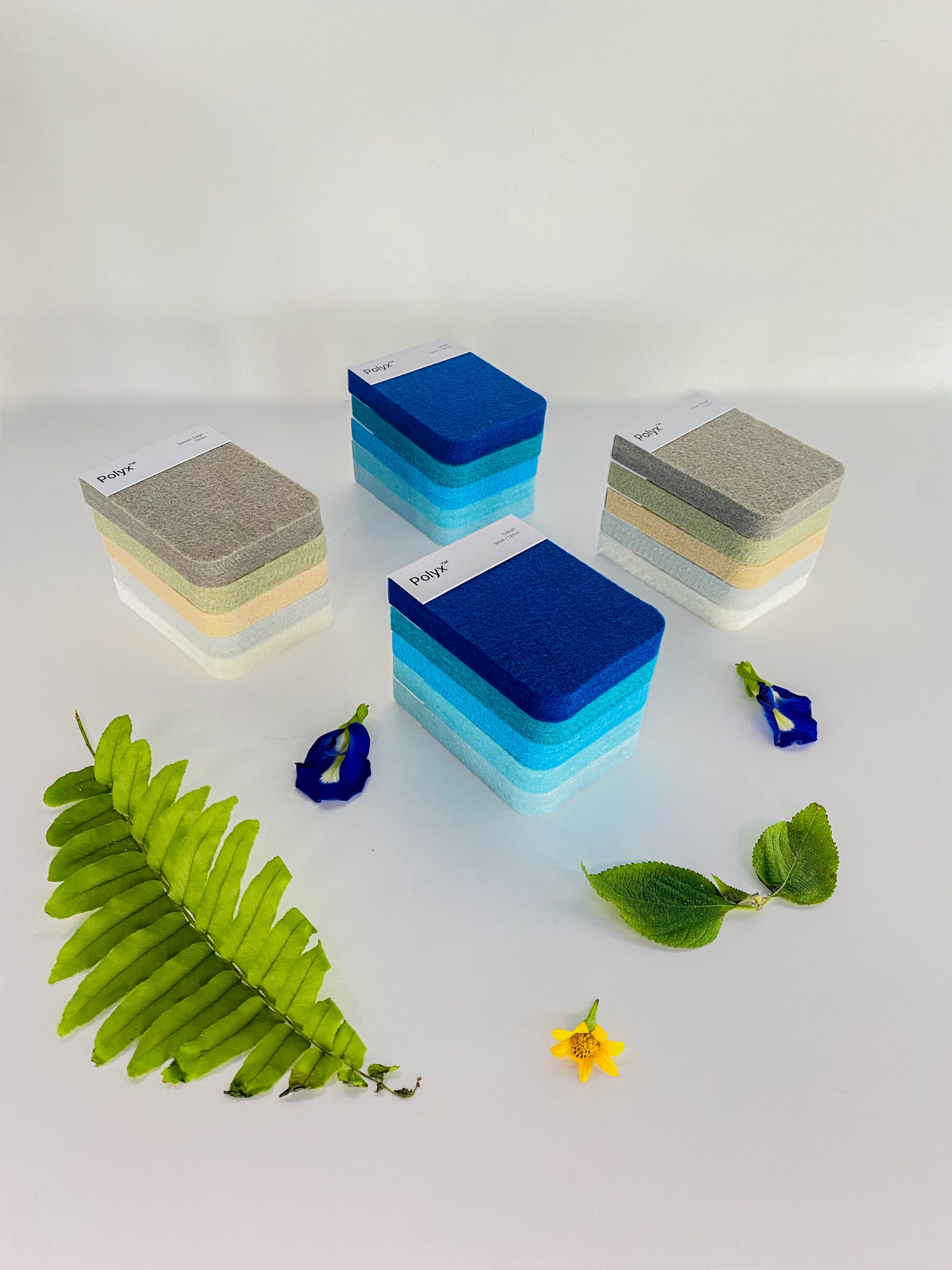 Stacked rectangular fabric samples in various shades of blue, grey, and beige sit on a white surface, with decorative green leaves and flowers placed around them.