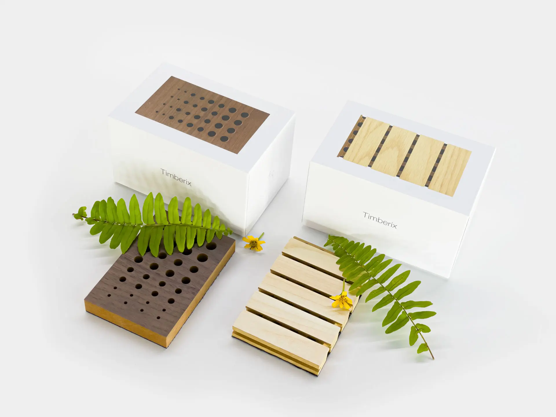 Two Timberix acoustic sample kits, one with perforated holes and the other with slotted grooves, displayed with green fern leaves and small yellow flowers for decoration.