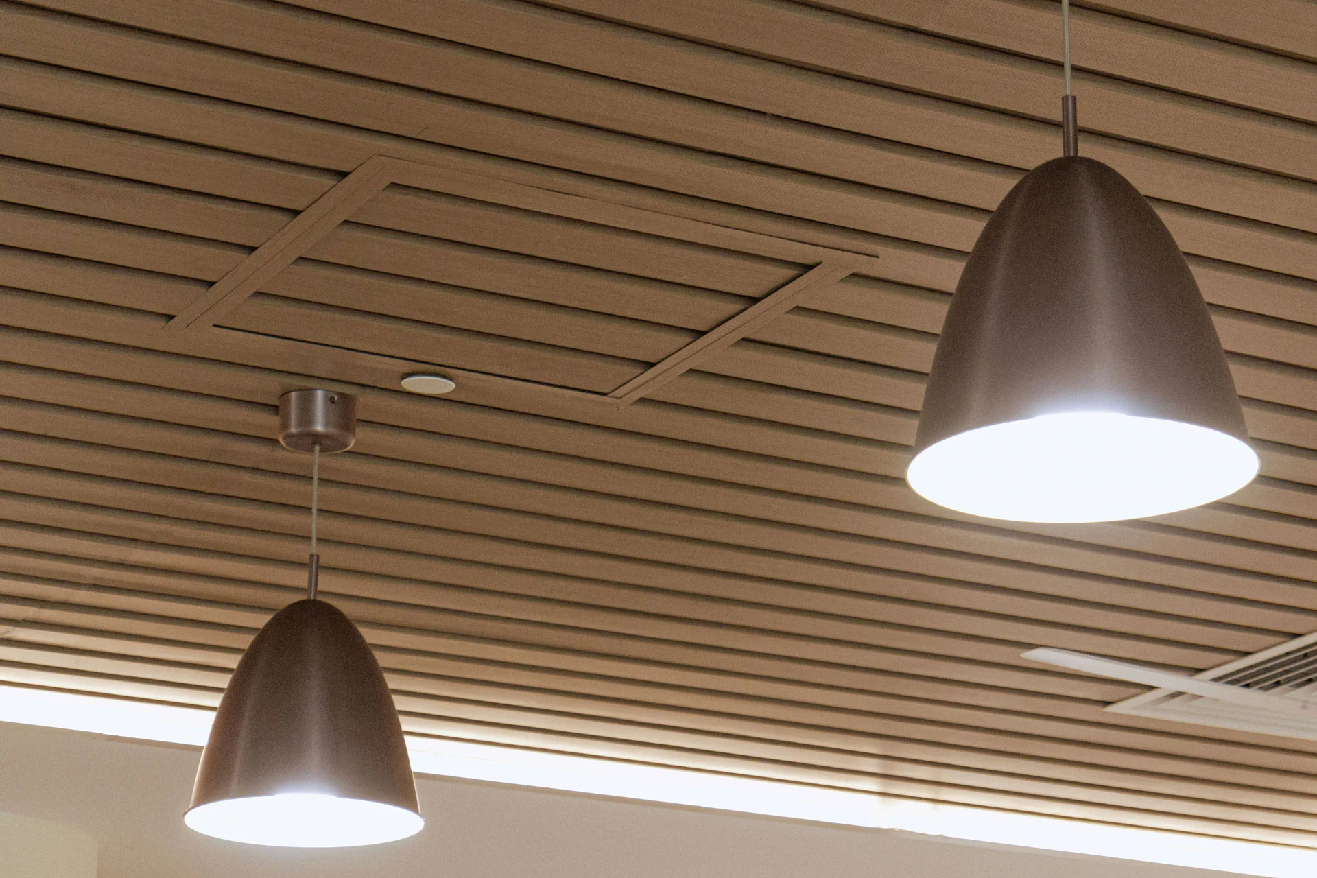 Two modern pendant lights hang from a wooden slatted ceiling, both turned on and emitting a soft white light. The ceiling has a hatch and an air vent visible among the slats.