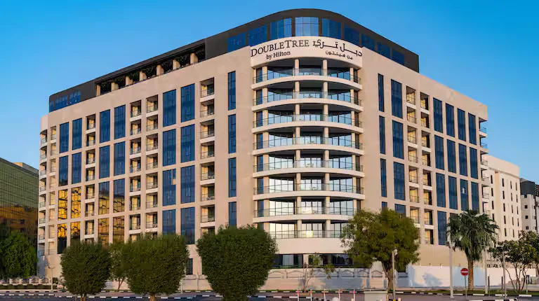 A modern multi-story hotel building with large windows and balconies, labeled DoubleTree by Hilton in English and Arabic. The hotel is surrounded by trees and set against a clear blue sky.