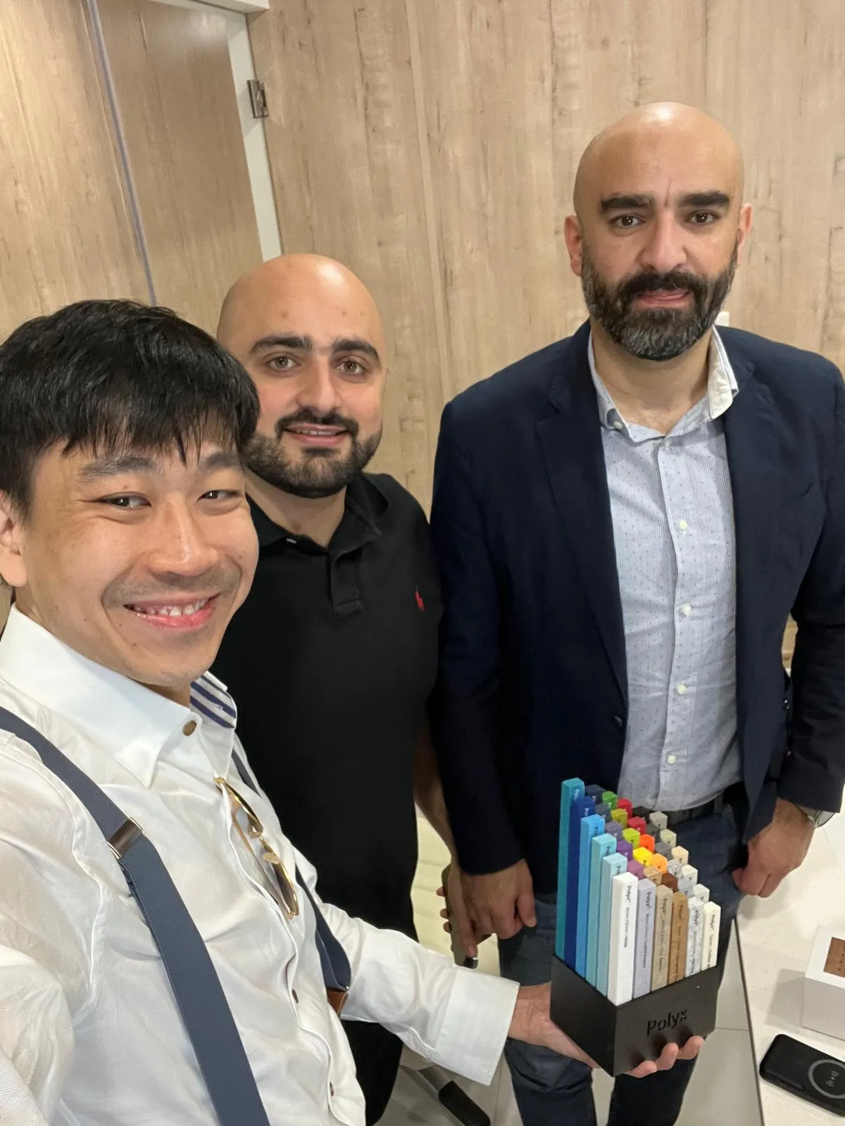 Three men stand together indoors, smiling at the camera. One holds a display of colorful rectangular samples arranged upright in a black stand. The background shows light wooden panels and a door.