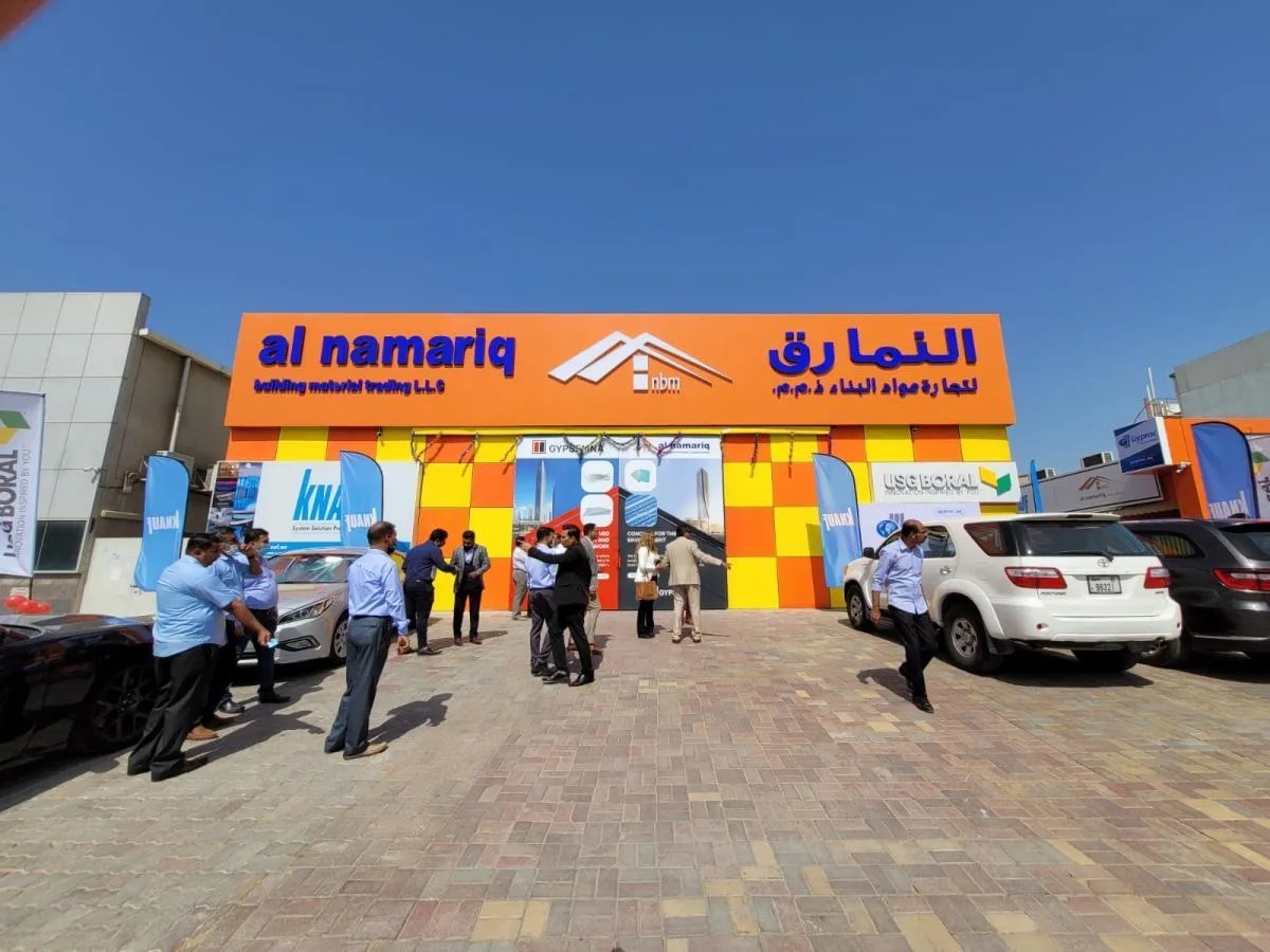 People stand and walk outside a bright orange and yellow building with the sign “al namariq” in English and Arabic. Several cars are parked in front under a clear blue sky.
