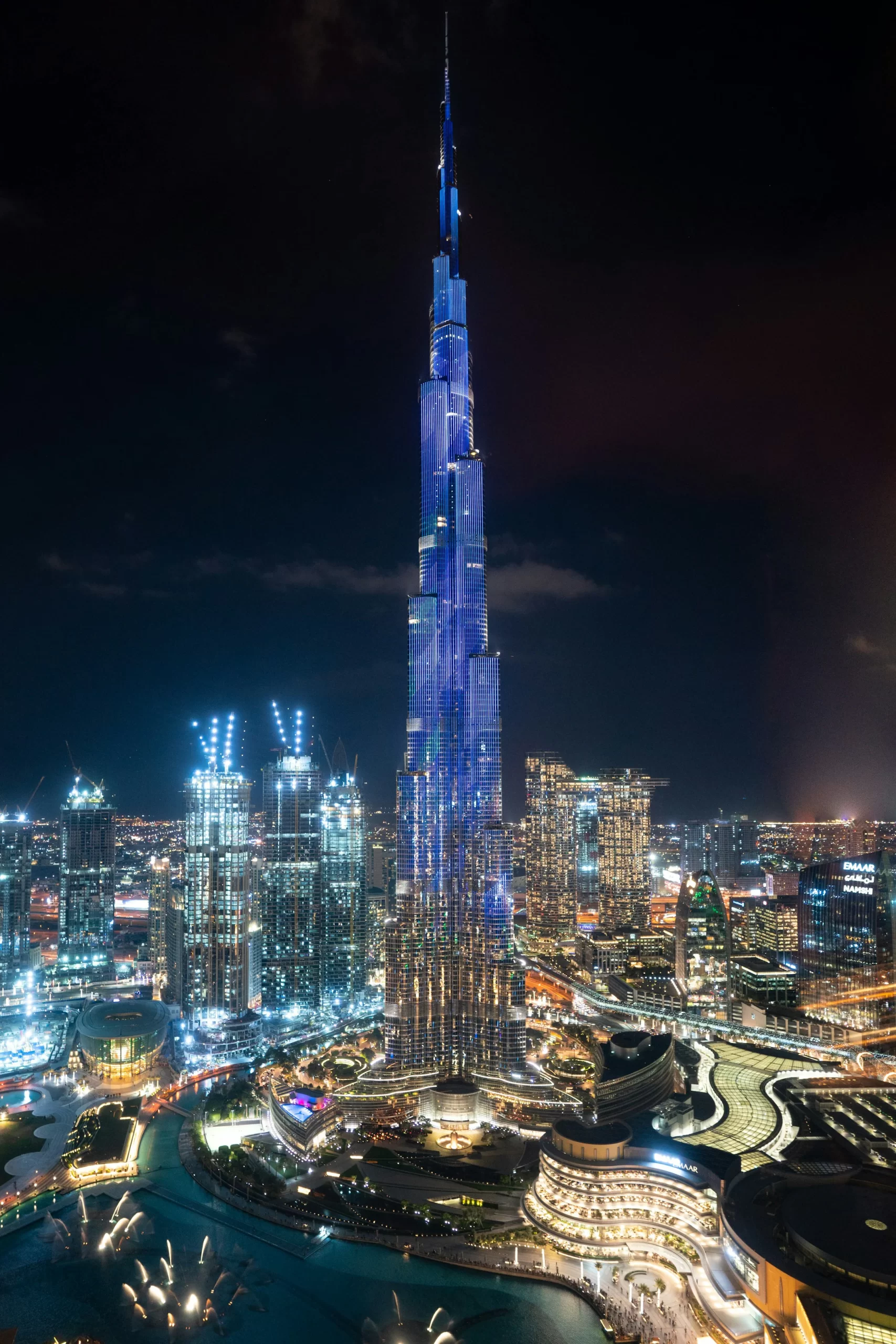 The Burj Khalifa in Dubai illuminated against the night sky, surrounded by other skyscrapers. The city lights reflect off the water in the foreground.