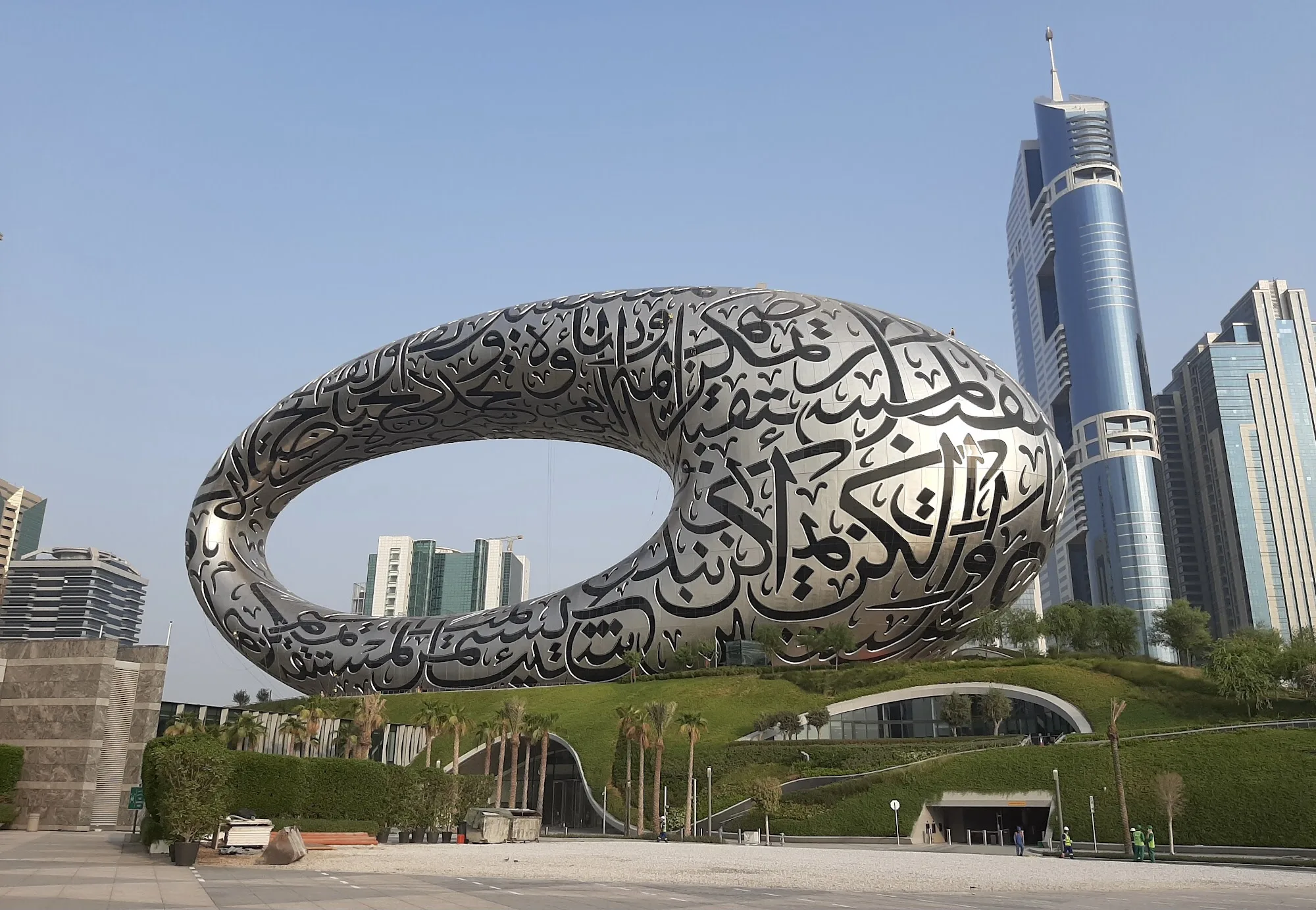 A large, futuristic silver building with Arabic calligraphy on its surface, shaped like an oval ring with an open center, stands in front of modern skyscrapers and green landscaping under a clear sky.