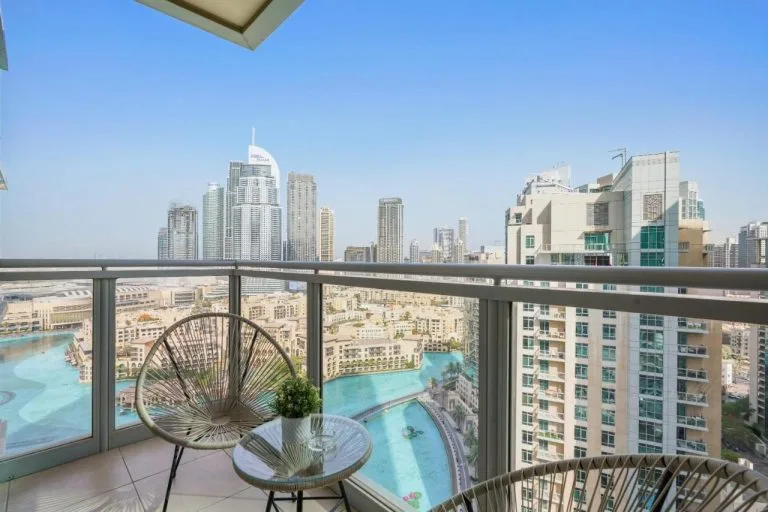 A balcony with two modern chairs and a small glass table overlooks a cityscape featuring tall skyscrapers and a large pool below, under a clear blue sky.