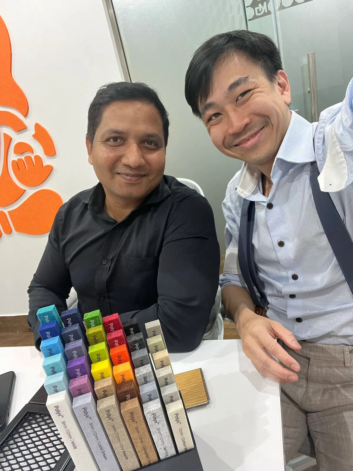 Two men smiling and sitting at a table with a display of colorful material samples in front of them. A bright orange wall art is visible in the background. The setting appears to be an office or meeting room.