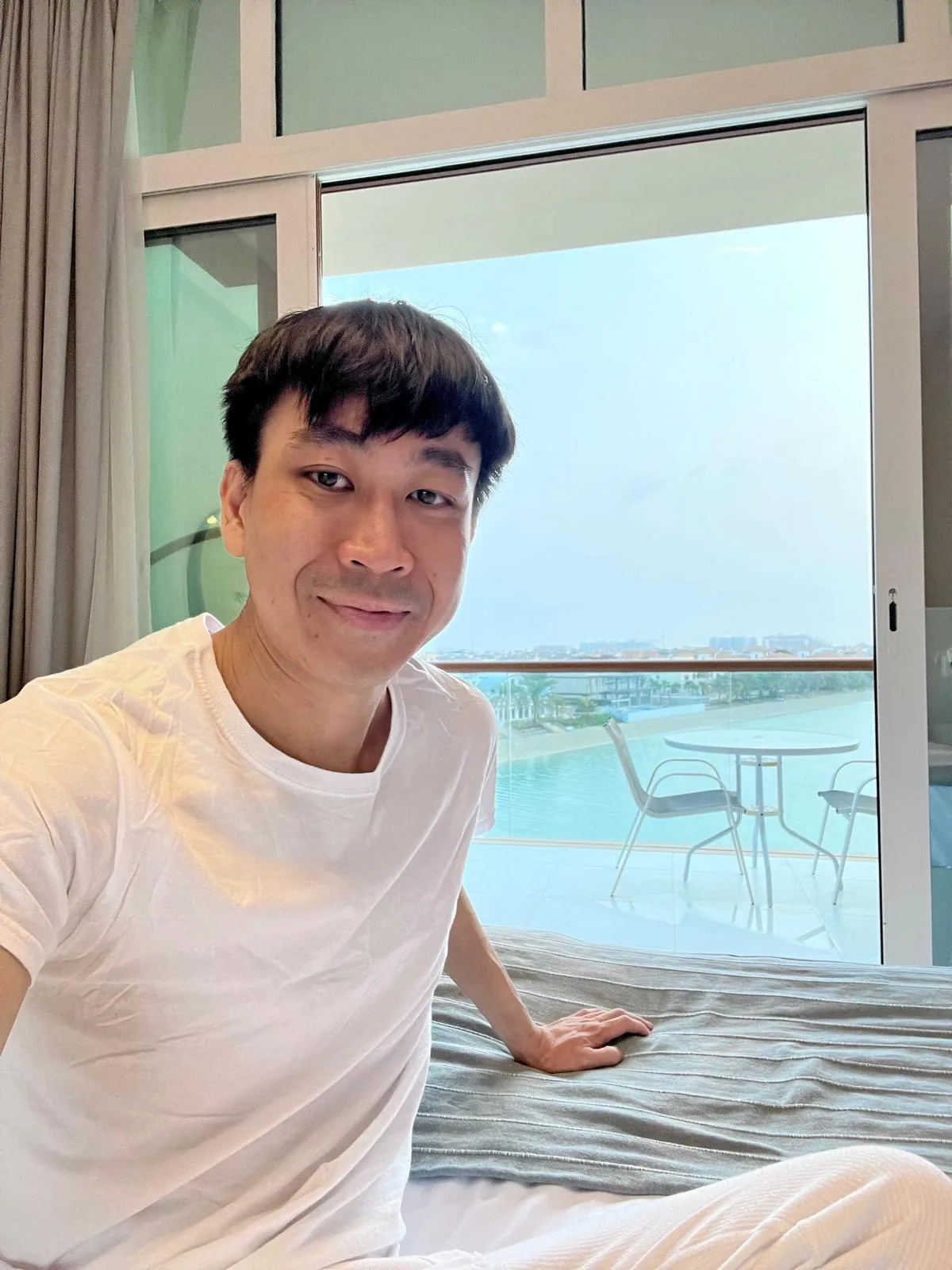 A person in a white shirt sits on a bed, smiling at the camera. Behind them is a large glass door leading to a balcony with chairs and a view of water and distant buildings.