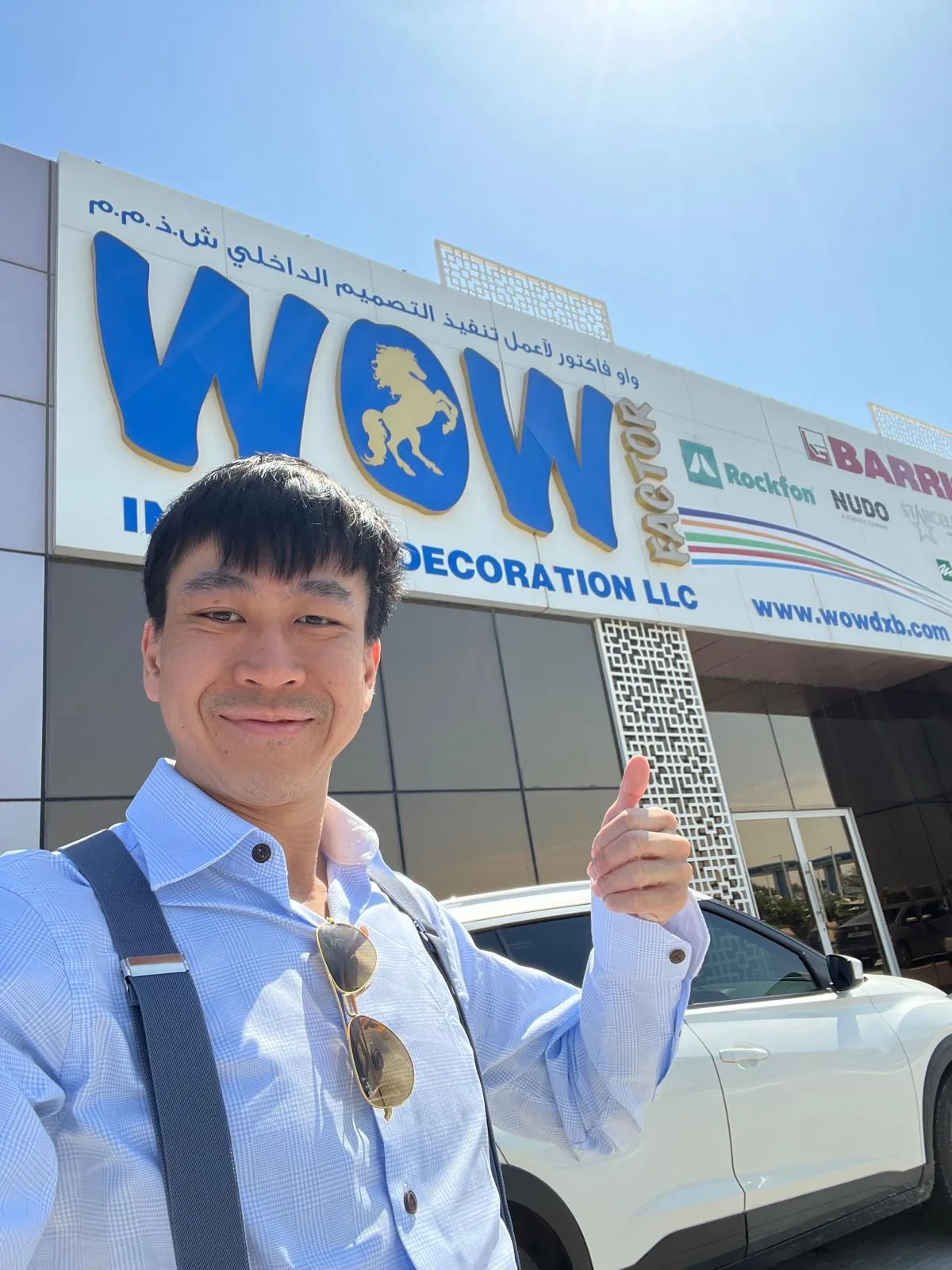 A man in a light blue shirt smiles and gives a thumbs-up outside the WOW Interior Decoration LLC building, with a white car parked in front and the company sign visible above in English and Arabic.