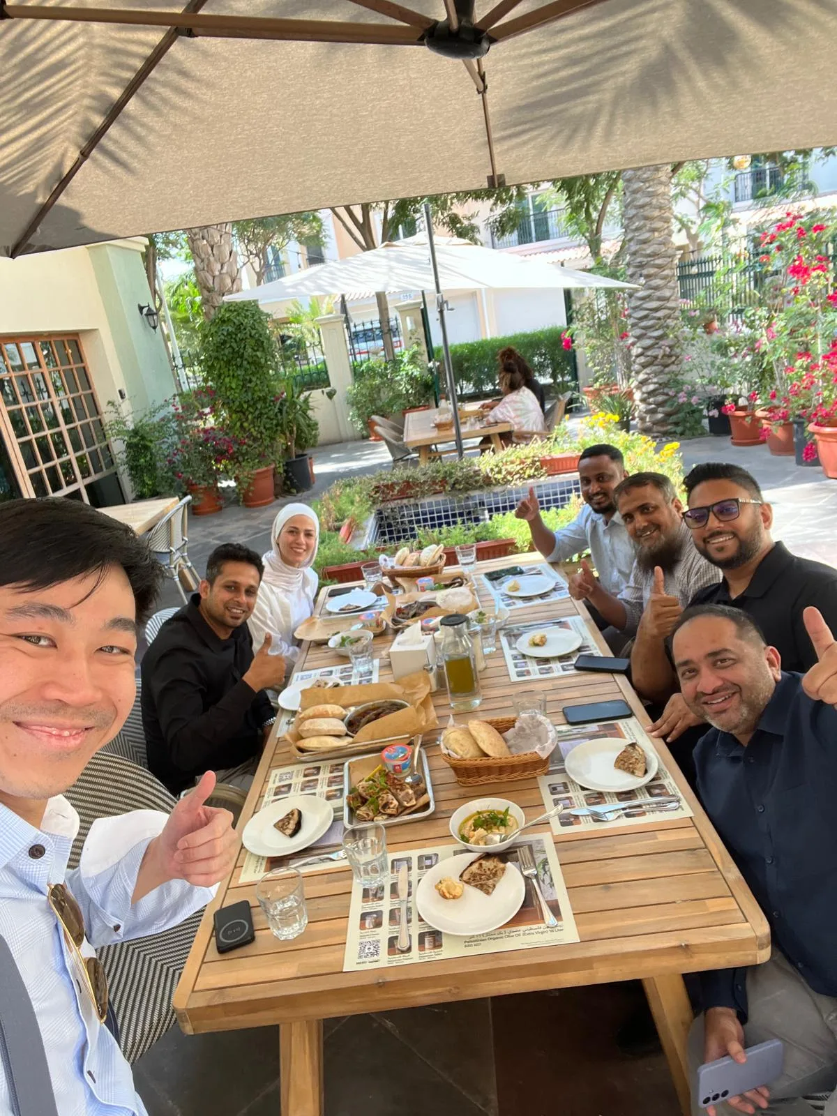A group of seven people sit around an outdoor wooden table covered with plates of food, smiling and giving thumbs up. They are under an umbrella, surrounded by greenery and a sunny, relaxed atmosphere.