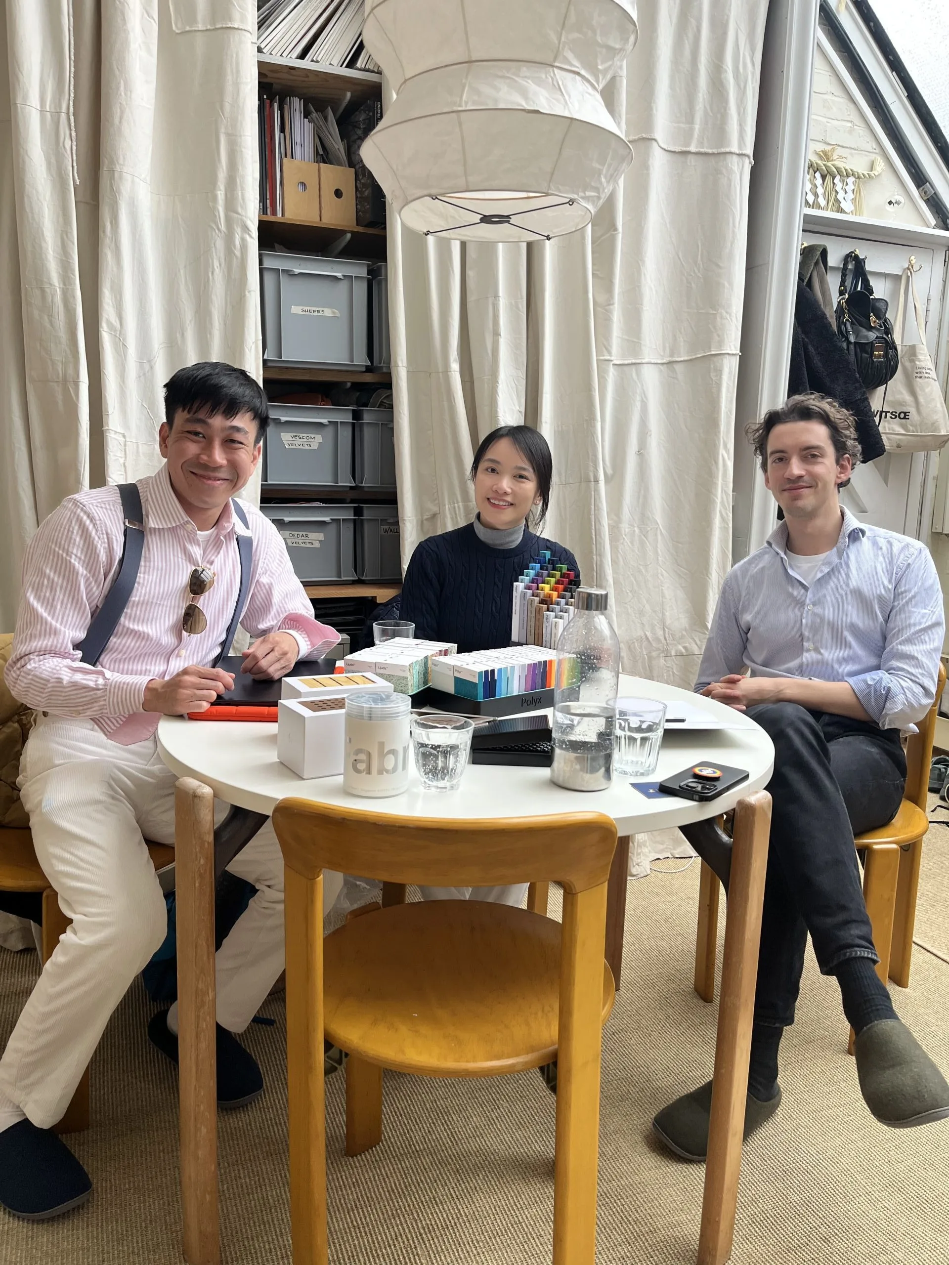 Three people sit around a round table with art supplies, notebooks, and water bottles, smiling at the camera in a cozy, well-lit room with shelves, storage boxes, and a curtain backdrop.