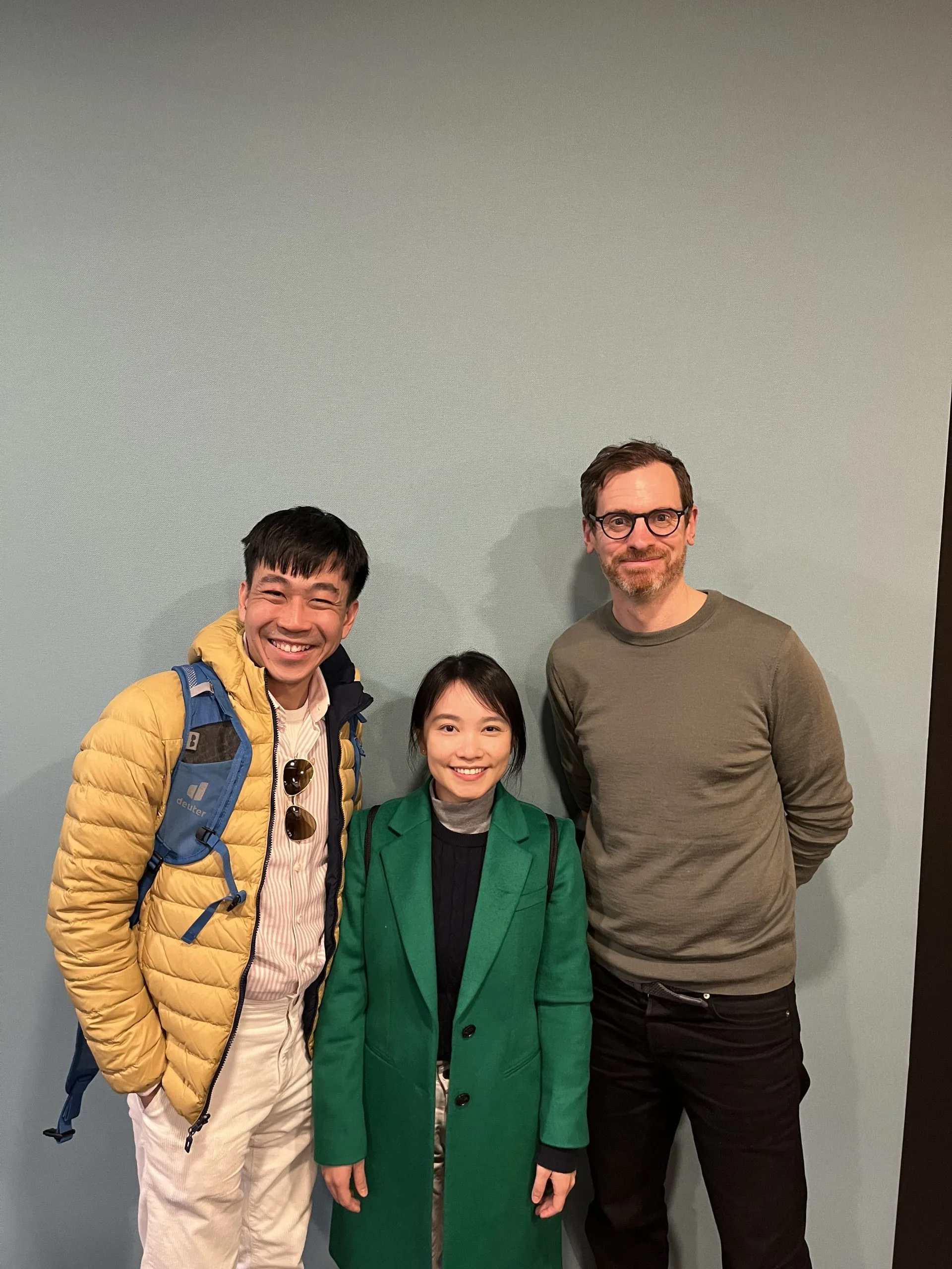 Three people stand smiling in front of a plain light blue wall. One person wears a yellow jacket and backpack, the middle person wears a green coat, and the third wears glasses and a green sweater.