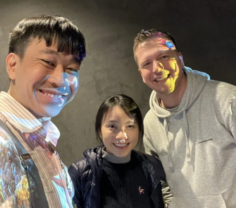 Three people are smiling and posing together indoors, standing close to each other. Colorful light reflections appear on their faces and clothing. The background is a plain, dark wall.