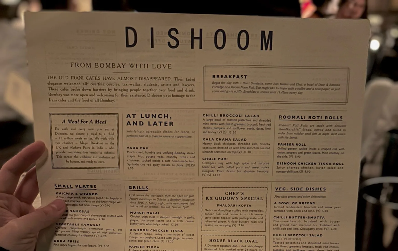 A hand holding a Dishoom restaurant menu, featuring sections like Breakfast, At Lunch, and Later, and listings for various Indian dishes, descriptions, and prices on a beige background. The setting appears dimly lit.