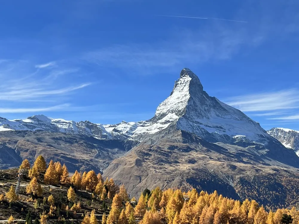 The image depicts the iconic Matterhorn mountain in Switzerland, standing tall with its jagged, snow-covered peak against a clear blue sky. Below the mountain, there are autumn-colored trees in shades of golden yellow, scattered across the rugged, rocky terrain, creating a contrast between the greenery and the snowy peaks in the background.