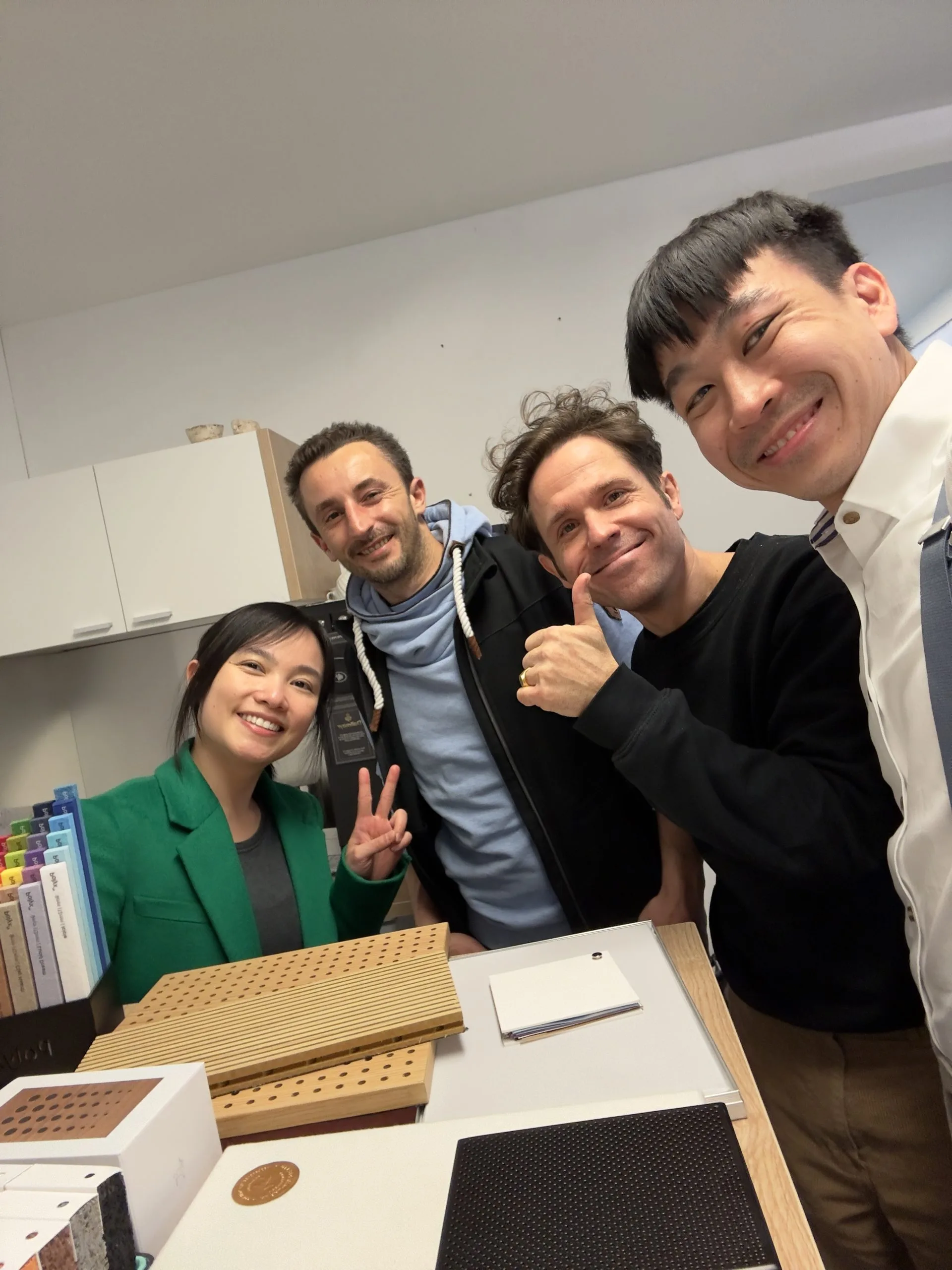 Four people stand together indoors, smiling at the camera. One woman in a green blazer flashes a peace sign, while the three men beside her are also smiling. Notebooks and swatches are on the desk in front of them.