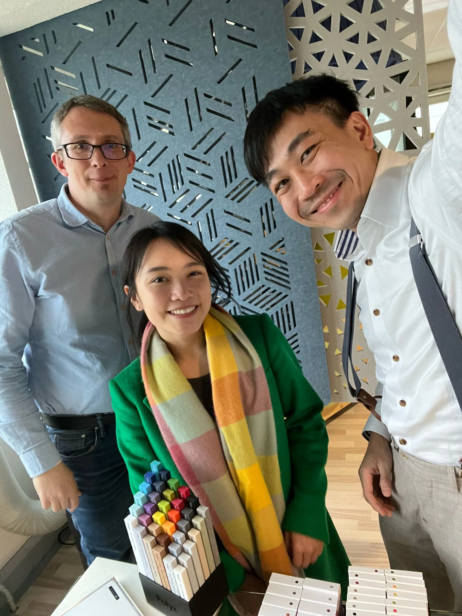 Three people smile at the camera in an indoor setting. One woman in a green coat and colorful scarf stands between two men, one in a blue shirt and glasses and the other in a white shirt with suspenders. Pens and notebooks are on the table.