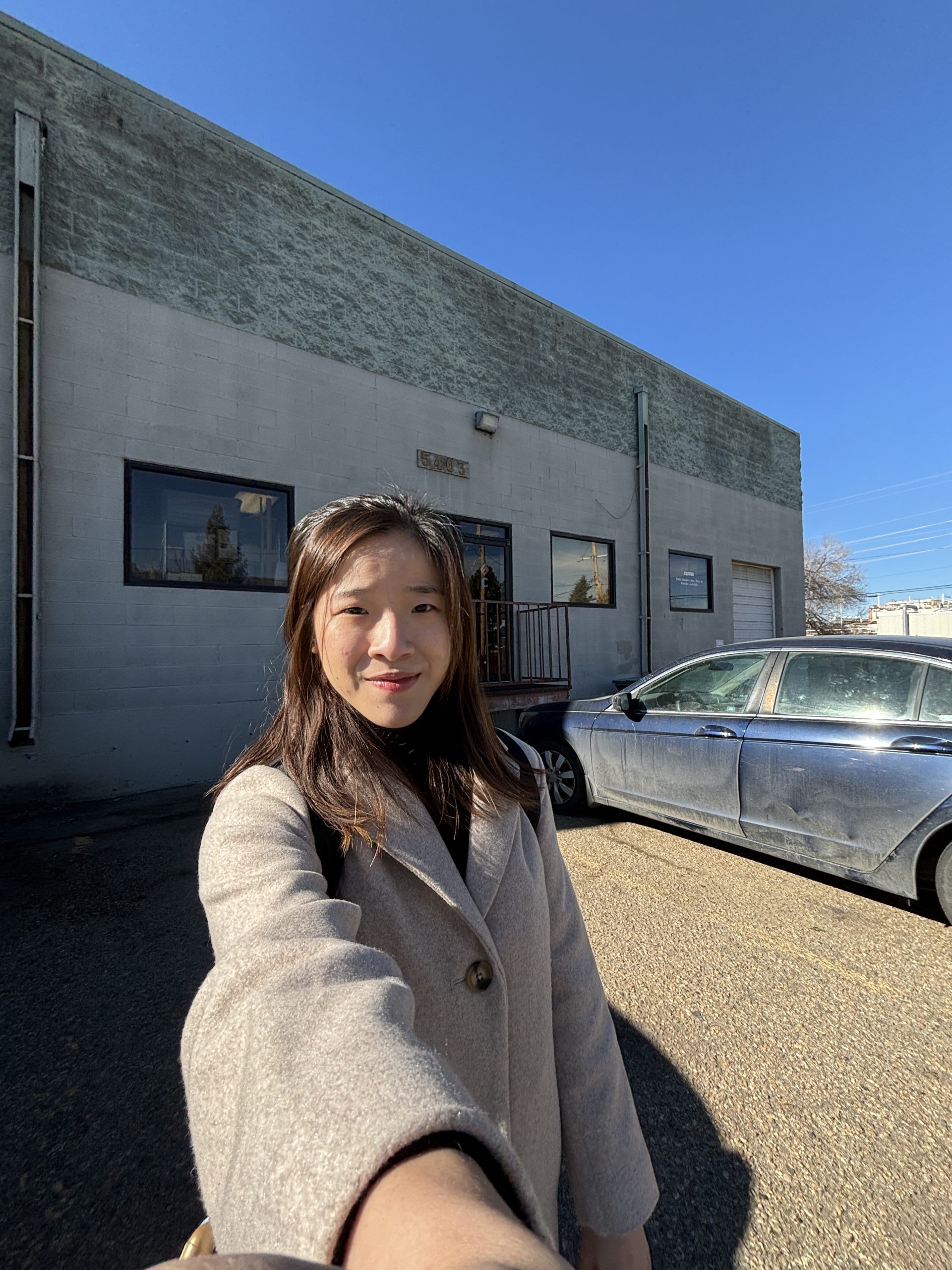 A woman taking a selfie outside a building under a clear blue sky, standing in front of a parked car.