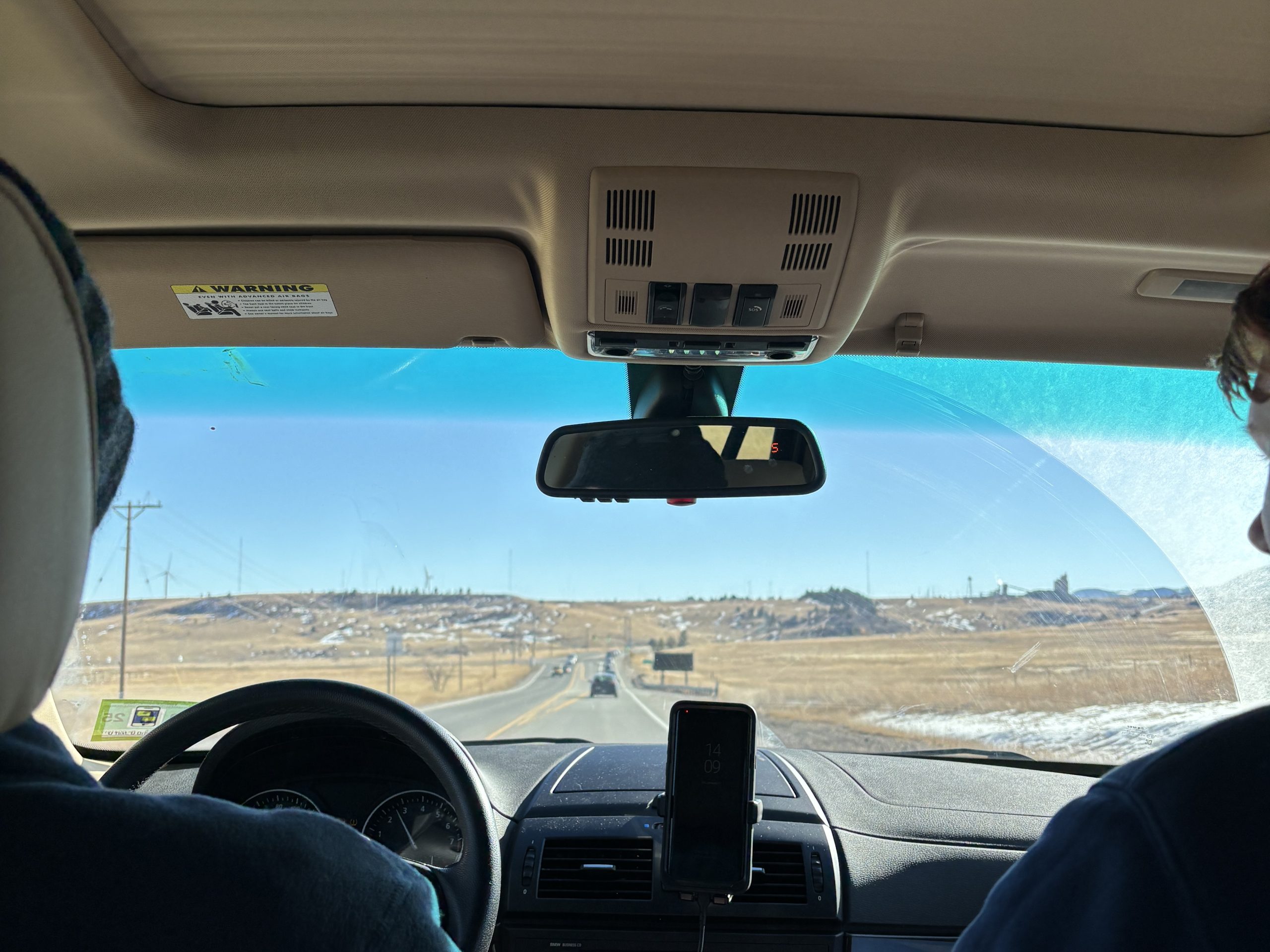 View from inside a car, looking out through the windshield at a clear blue sky and open road, with two people in the front seats driving through a rural landscape.