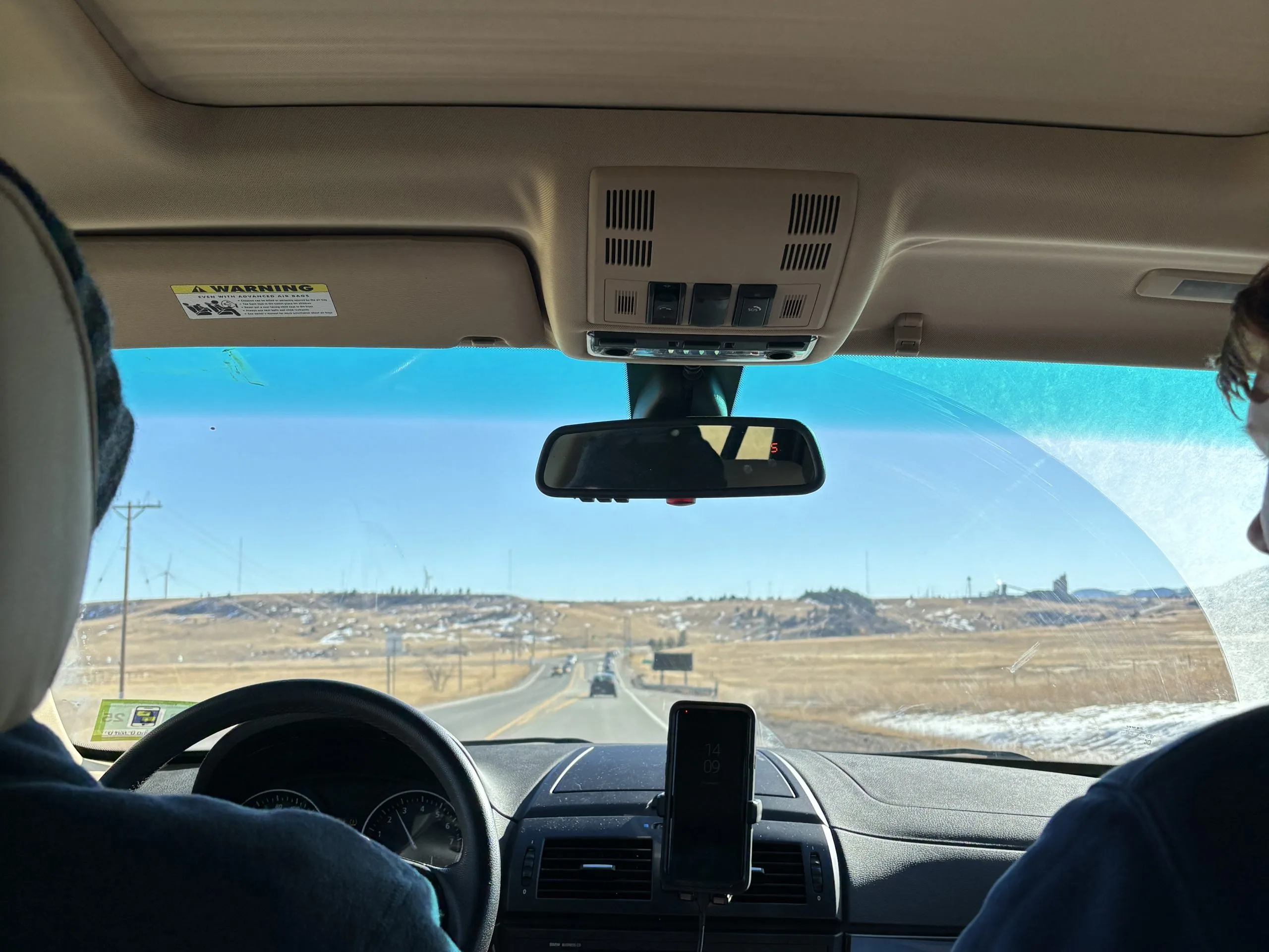 View from inside a car, looking out through the windshield at a clear blue sky and open road, with two people in the front seats driving through a rural landscape.