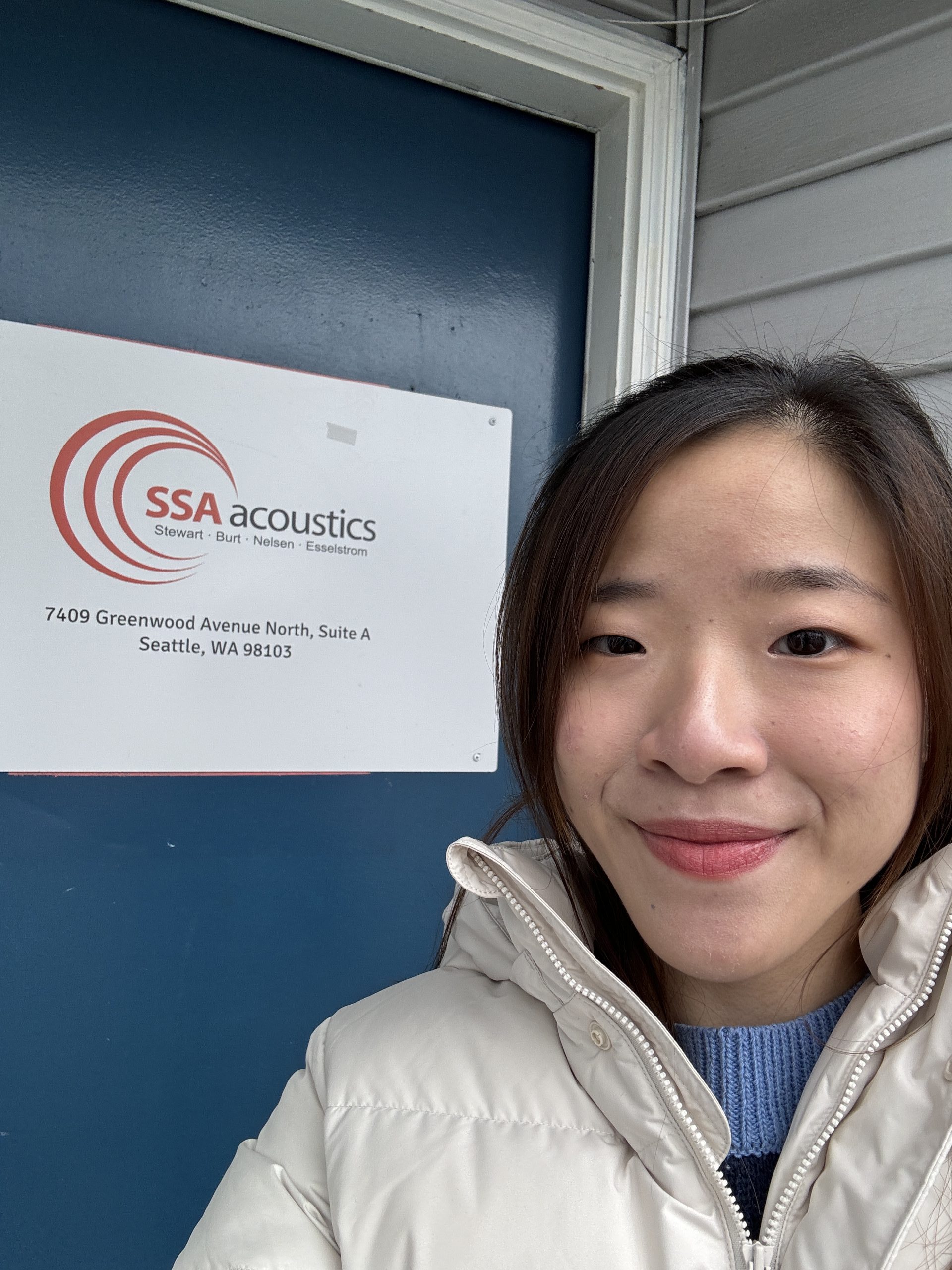 A selfie in front of a blue door, with a sign for SSA Acoustics visible, featuring a person smiling while wearing a warm, light-colored coat.