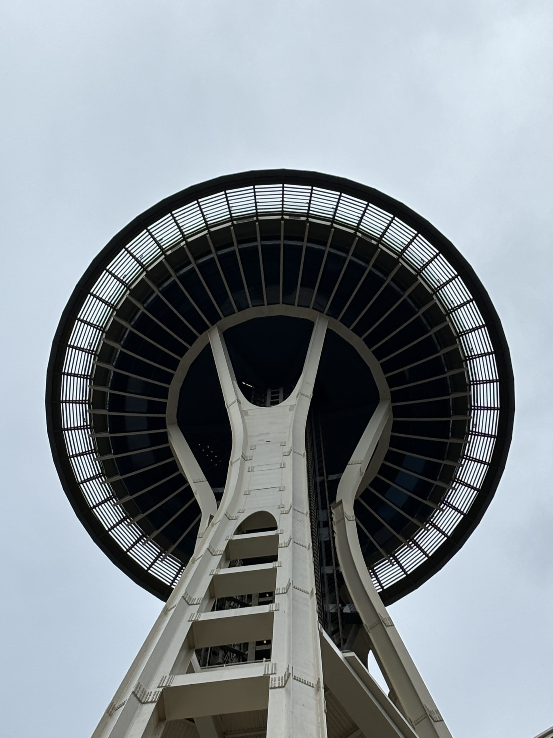 A close-up shot looking up at the iconic Space Needle from below, focusing on the tower's underside against a cloudy sky.