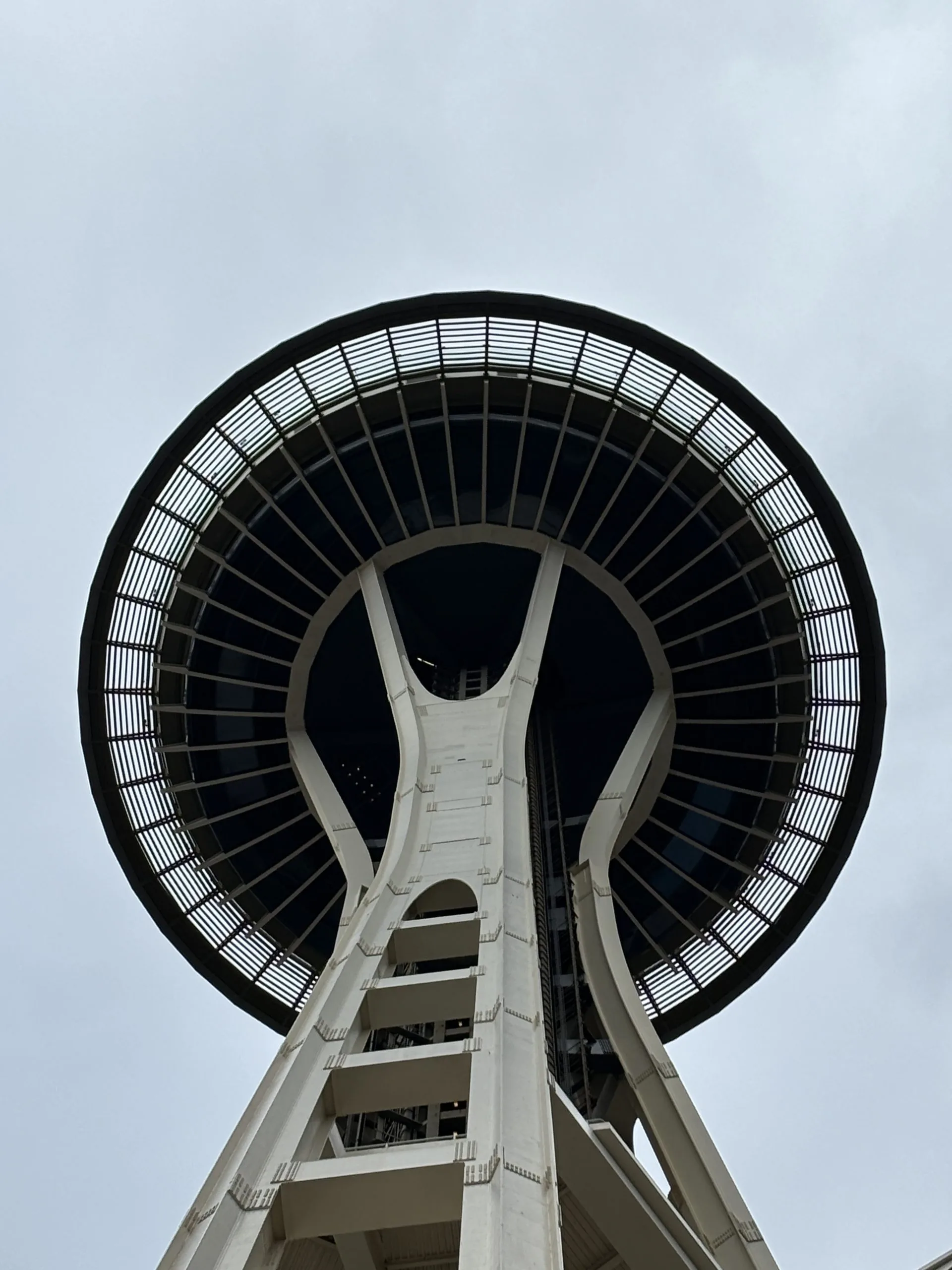 A close-up shot looking up at the iconic Space Needle from below, focusing on the tower's underside against a cloudy sky.
