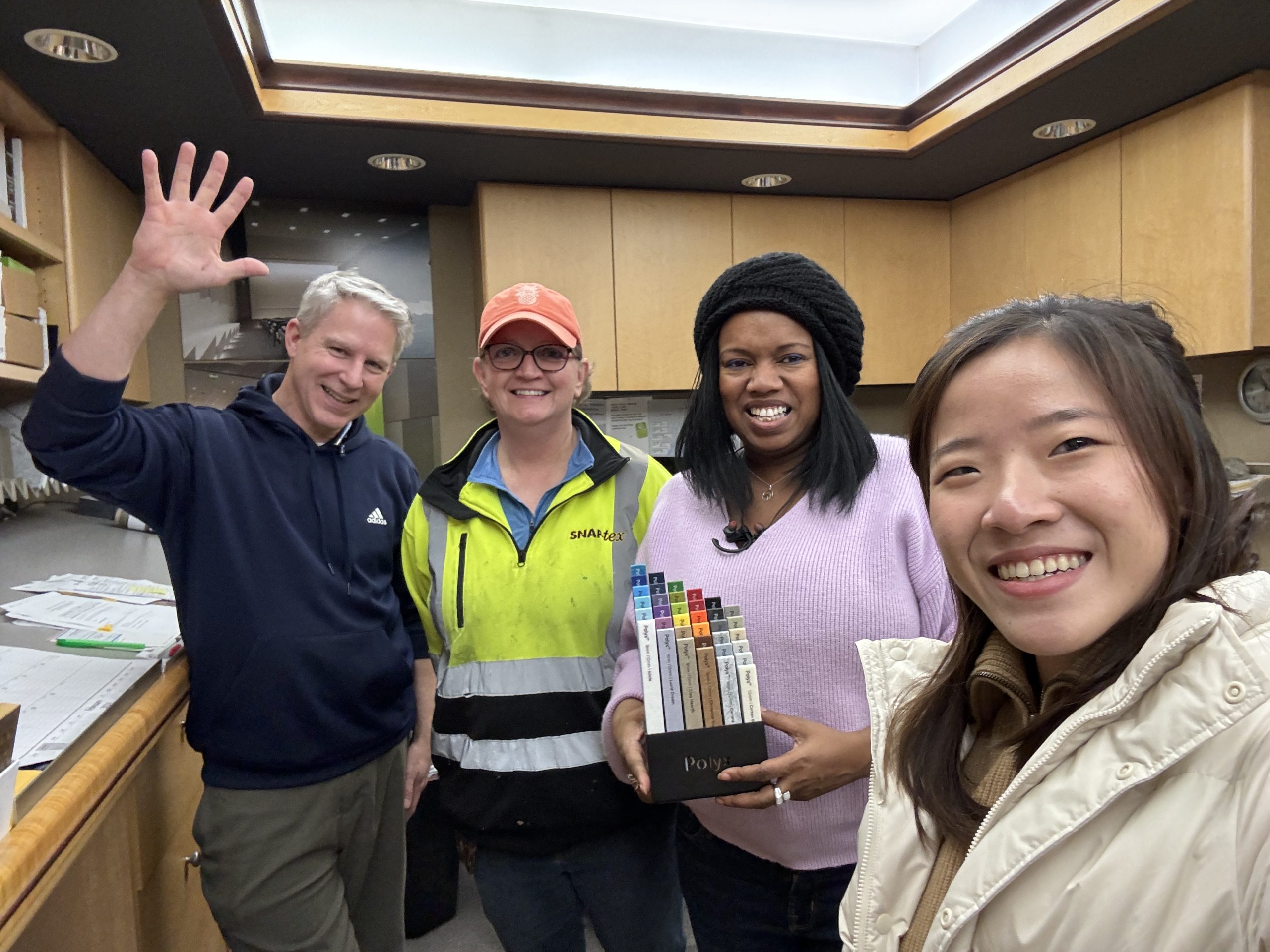 A group photo with four people smiling in an office environment, one person waving, another holding color samples.