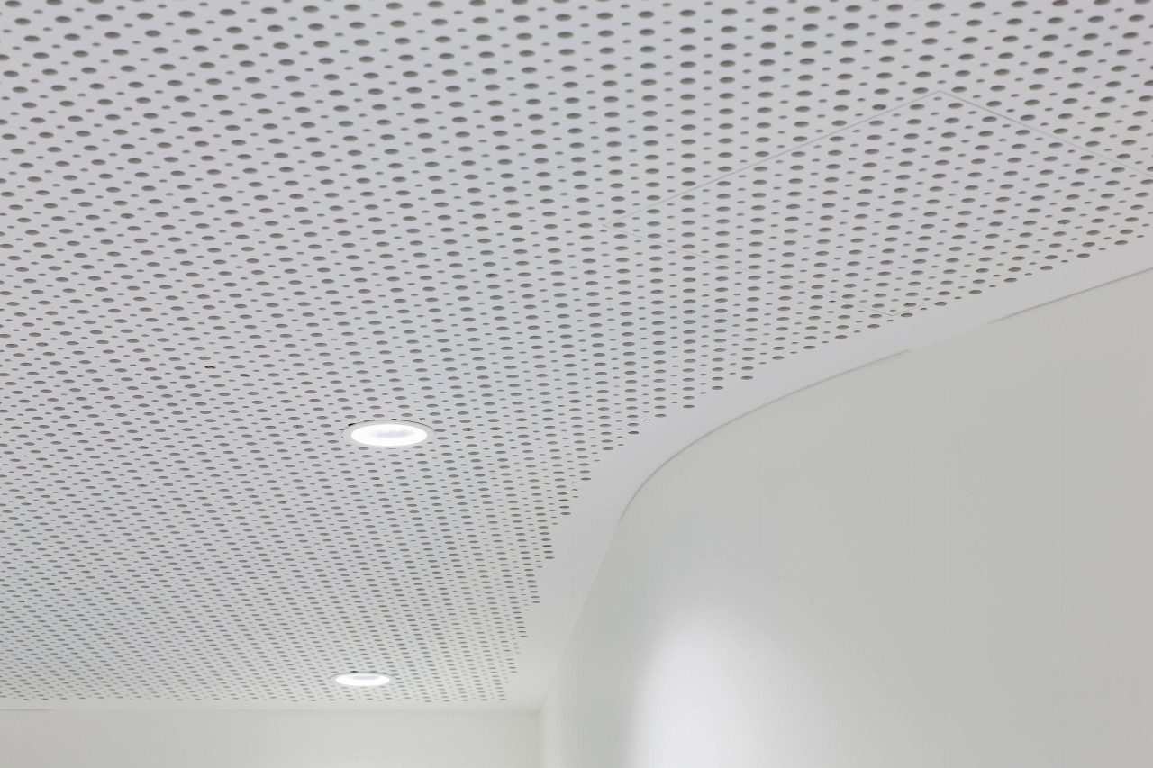 A white perforated ceiling with two recessed round lights and a curved wall below. The ceiling features a pattern of small, evenly spaced holes. The space appears modern and minimalistic.