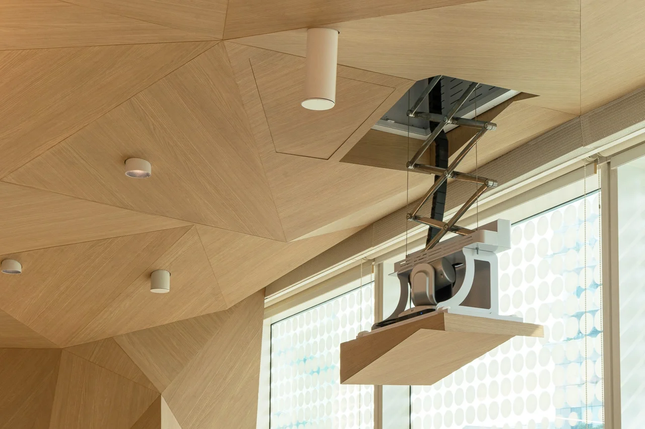 Modern wooden ceiling with geometric panels and a retractable mechanism attached to a tech device, in a clean, contemporary setting.
