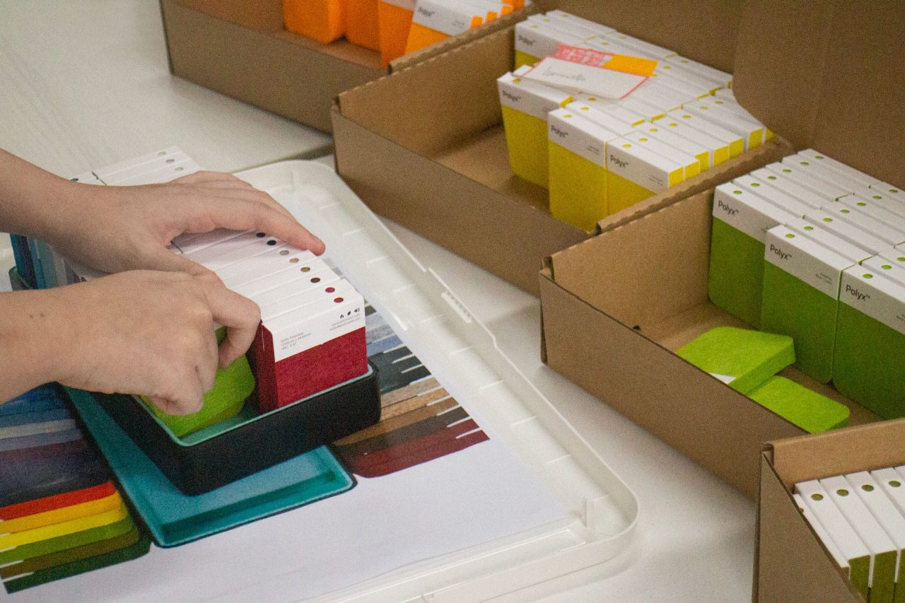 Hands organizing and placing rectangular sample products into a box, with various brightly colored samples visible in the background.