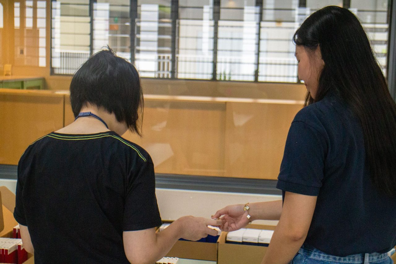 Two women standing by boxes filled with rectangular sample products, with one woman offering something to the other.