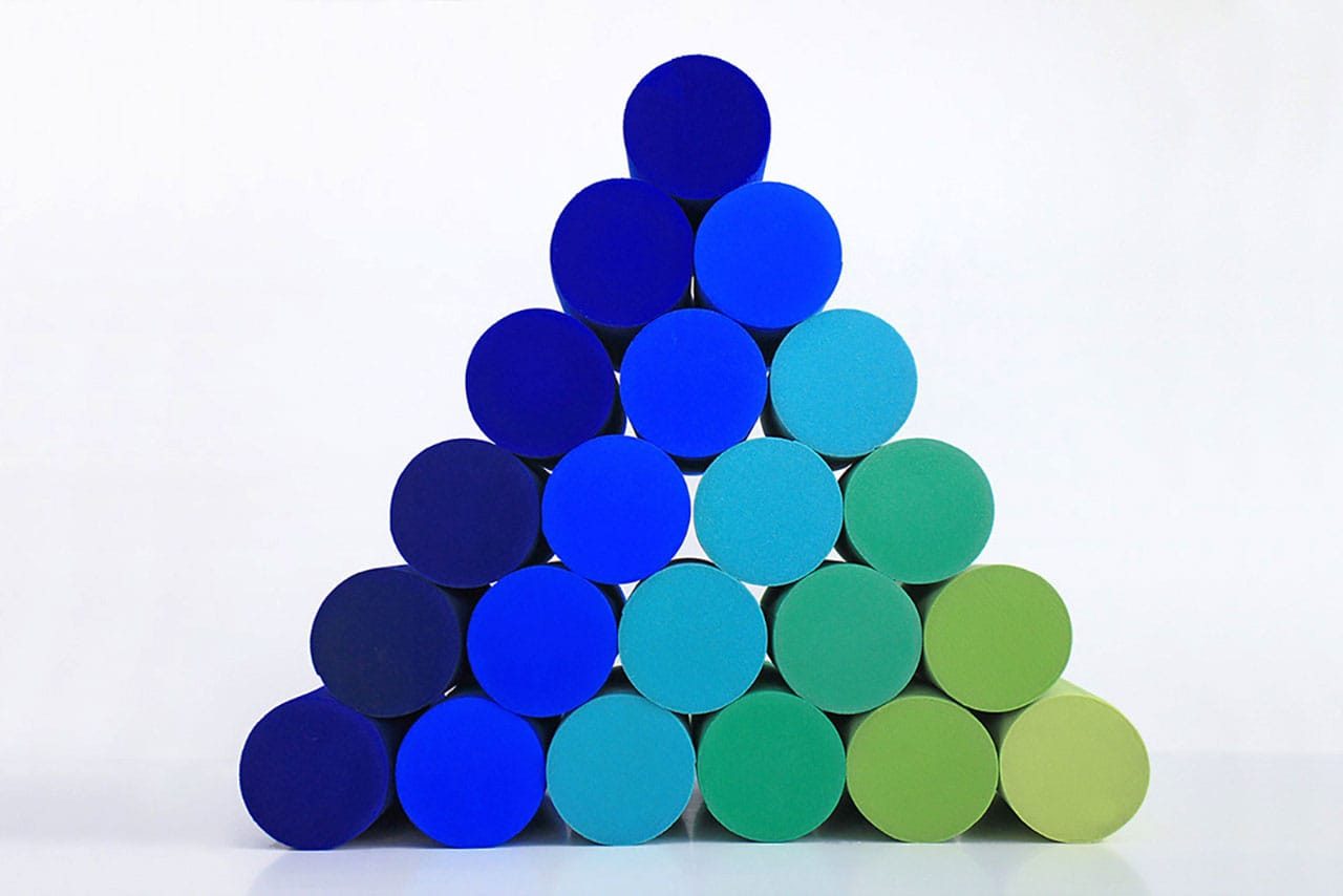 A pyramid made of cylindrical blocks in shades of blue and green is stacked against a white background. The colors transition from dark blue at the base to bright blue and green at the top and sides.