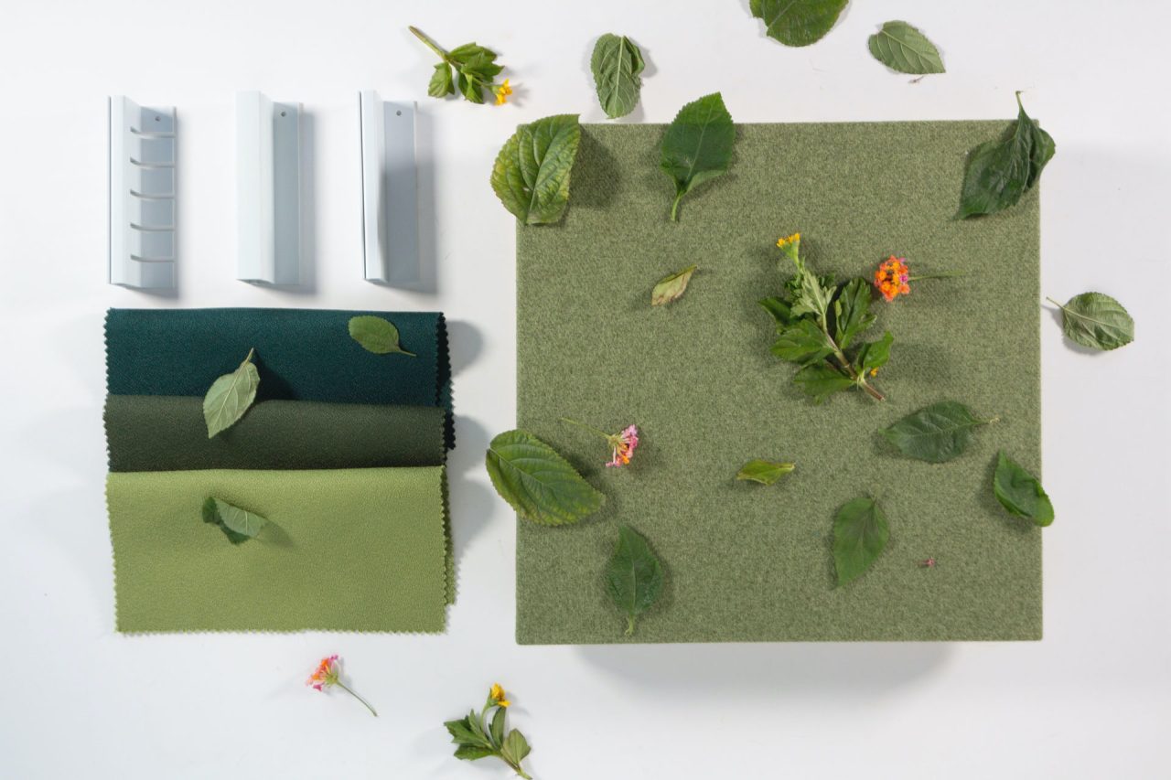 A green fabric square and fabric swatches in dark green, olive, and light green lie on a white surface, surrounded by scattered leaves, small flowers, and three gray plastic wall corner pieces.