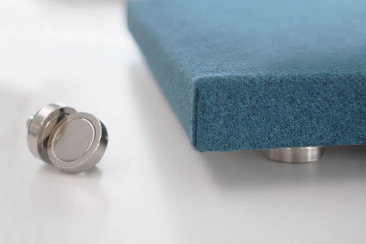 A close-up of a blue felt-like square panel supported by a metallic stand, with another round metallic element placed next to it, against a white background.