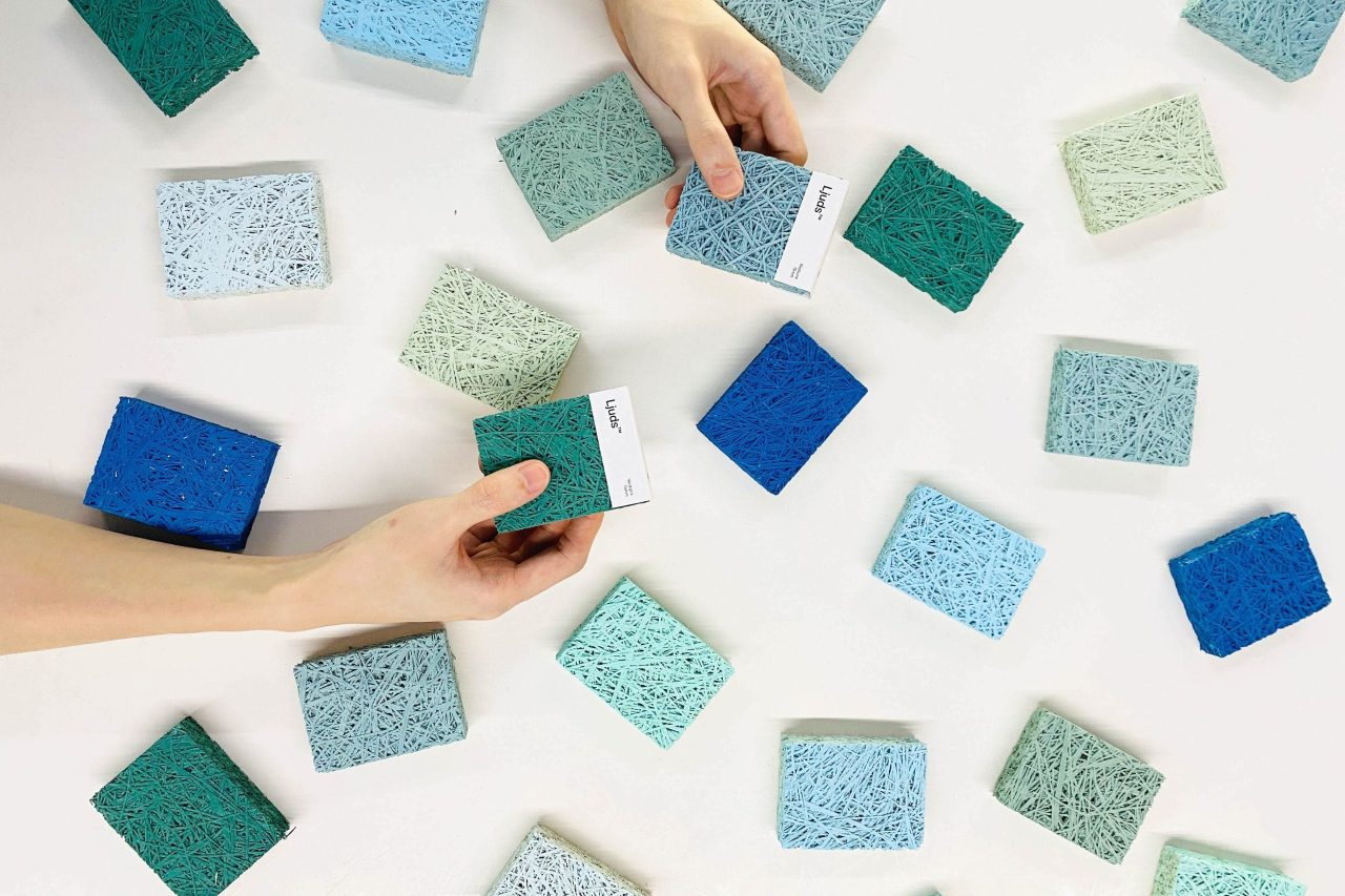A close-up view of two hands selecting from multiple textured sample blocks in shades of blue and green. The blocks are placed randomly on a white surface.