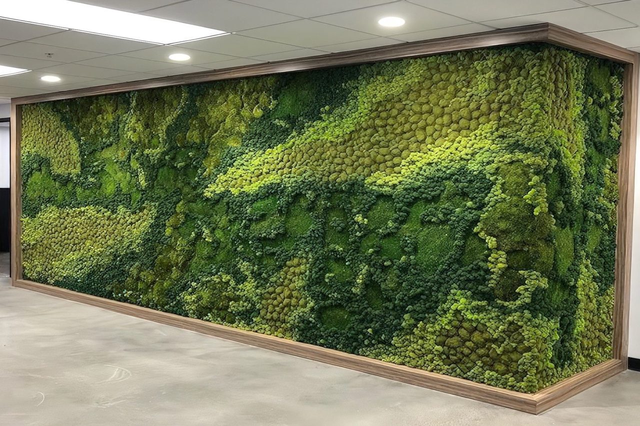 A large indoor wall covered with green and yellow preserved moss in organic patterns, framed with wood trim, under ceiling lights, on a polished concrete floor.