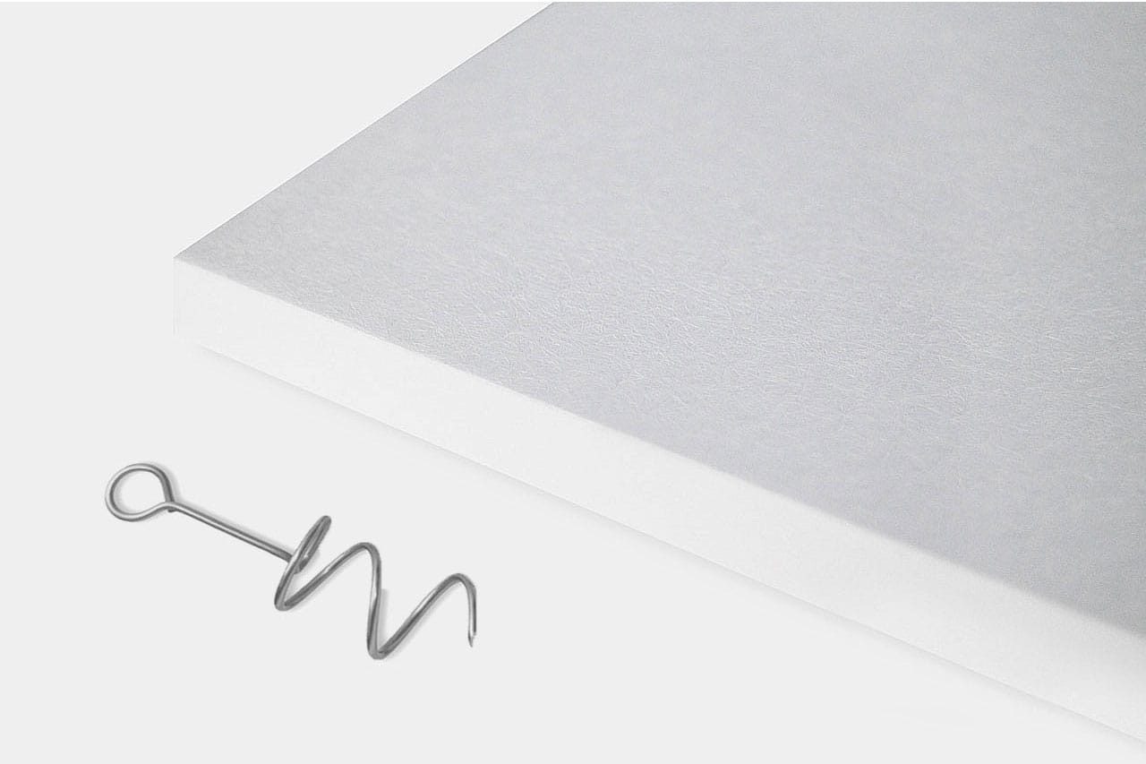 A thick, white rectangular sheet lies on a light surface next to a small, silver, spiral-shaped metal hook with a loop at one end.